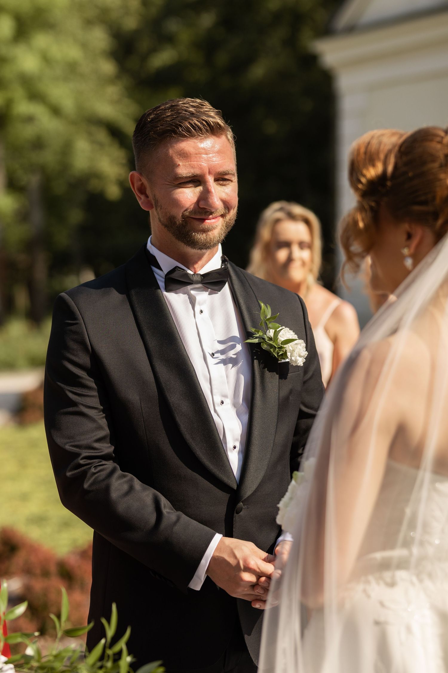 A groom in a black tuxedo shares an emotional moment with his bride during an outdoor wedding ceremony.
