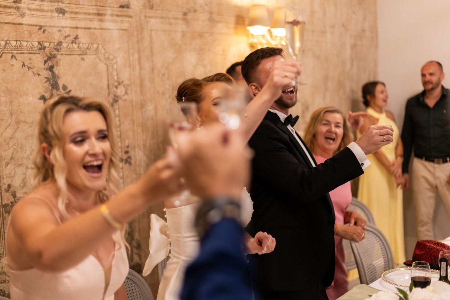 Wedding guests celebrate with joyful cheers and raised hands during a reception celebration.