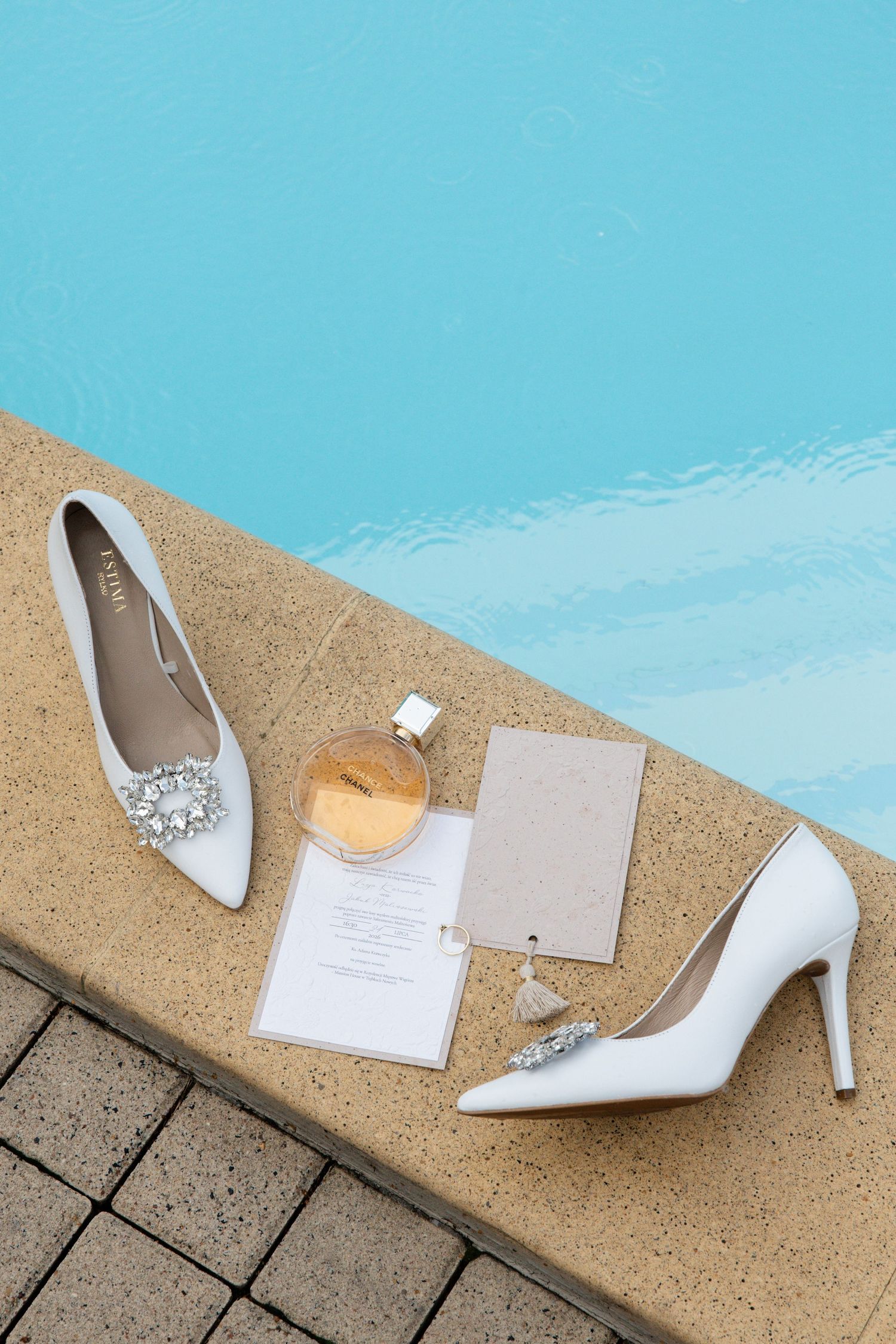 White bridal heels with crystal embellishment, wedding stationery, and perfume bottle arranged by a pool.