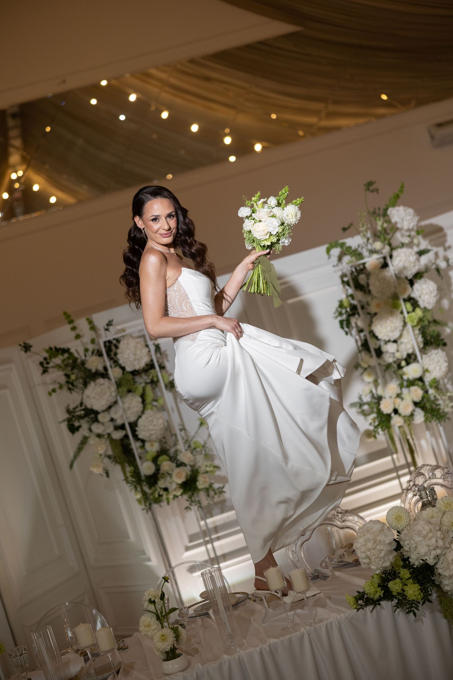 Elegant bride in white gown holding bouquet on swing, surrounded by white floral arrangements in decorated wedding venue.