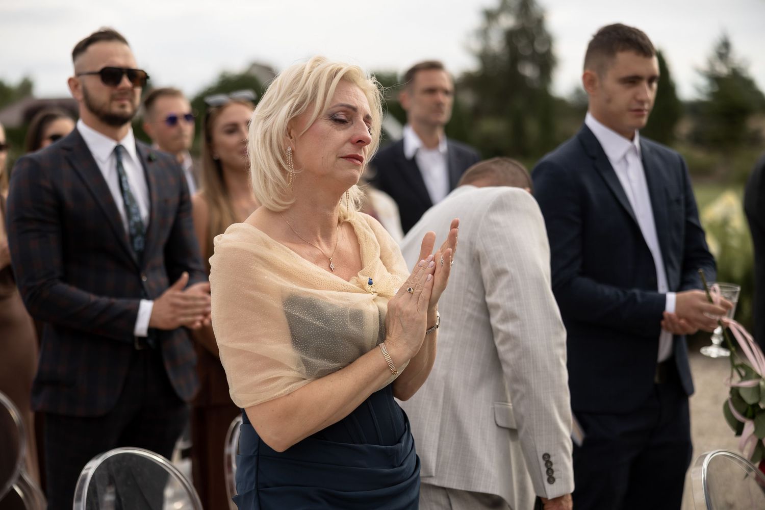 Emotional woman clapping hands at an outdoor wedding ceremony surrounded by guests in formal attire.