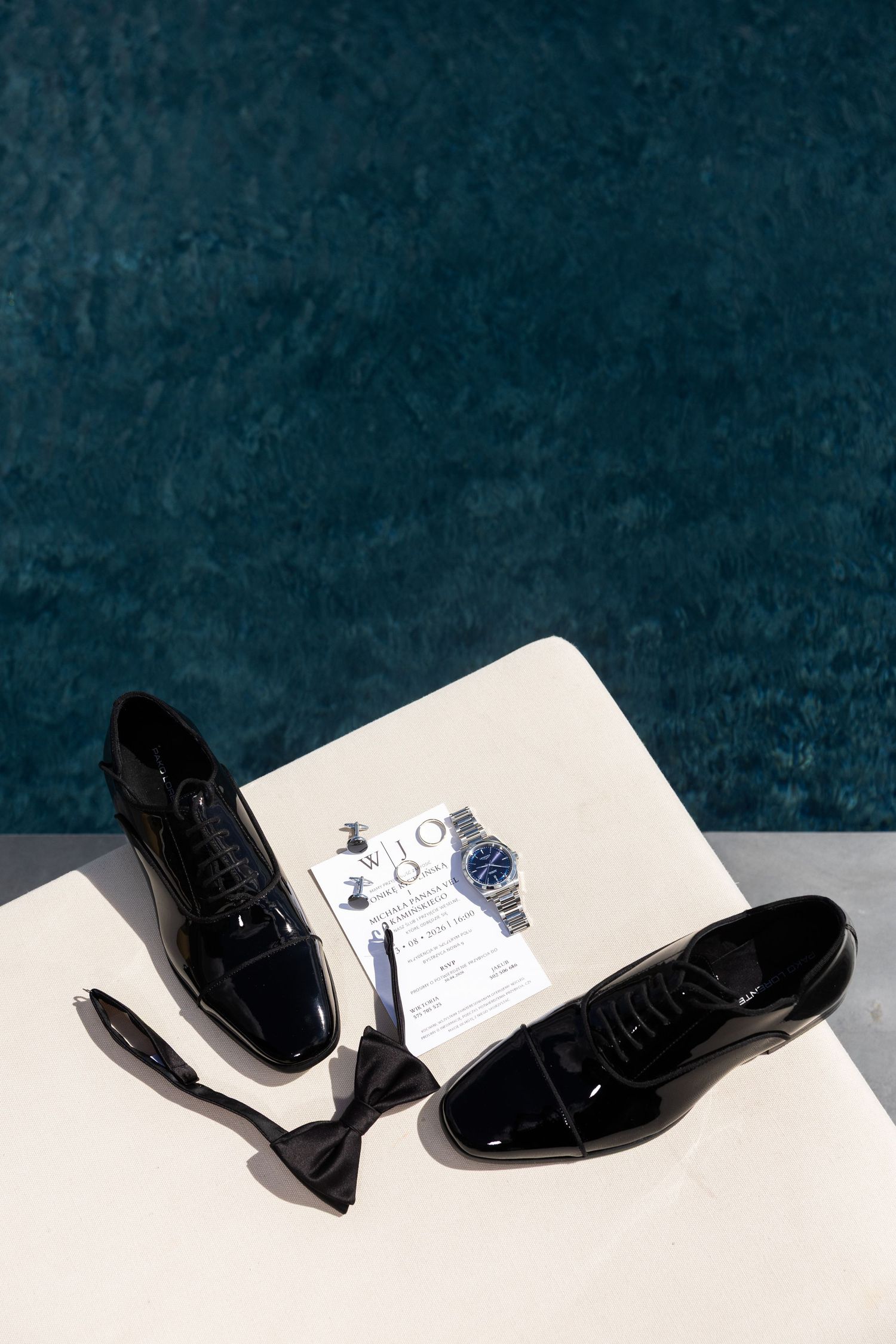 Black patent leather dress shoes, bow tie, watch, and cufflinks arranged on white surface for a formal wedding.