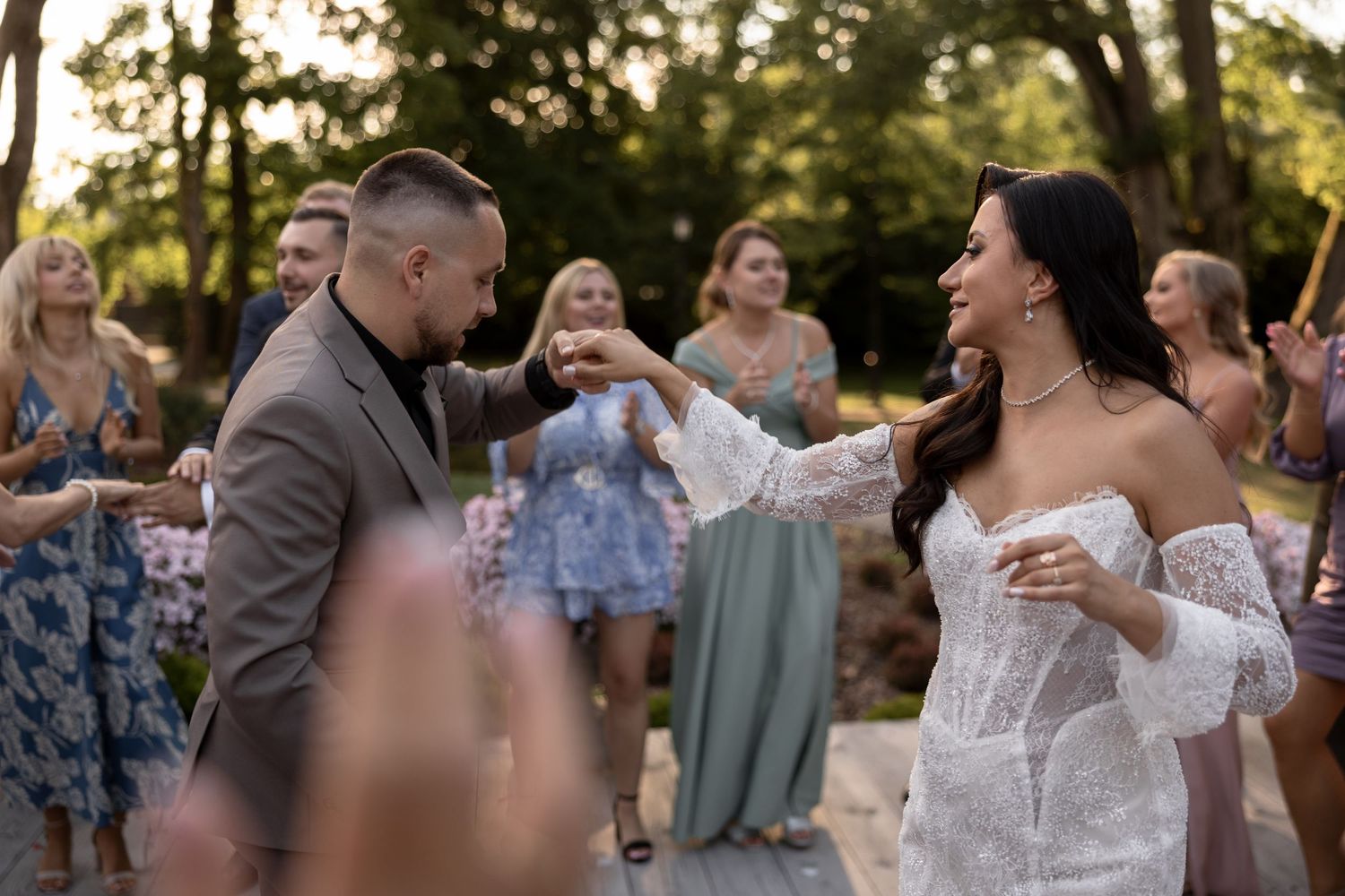 Bride and groom dancing joyfully outdoors at wedding reception surrounded by smiling guests in elegant attire.