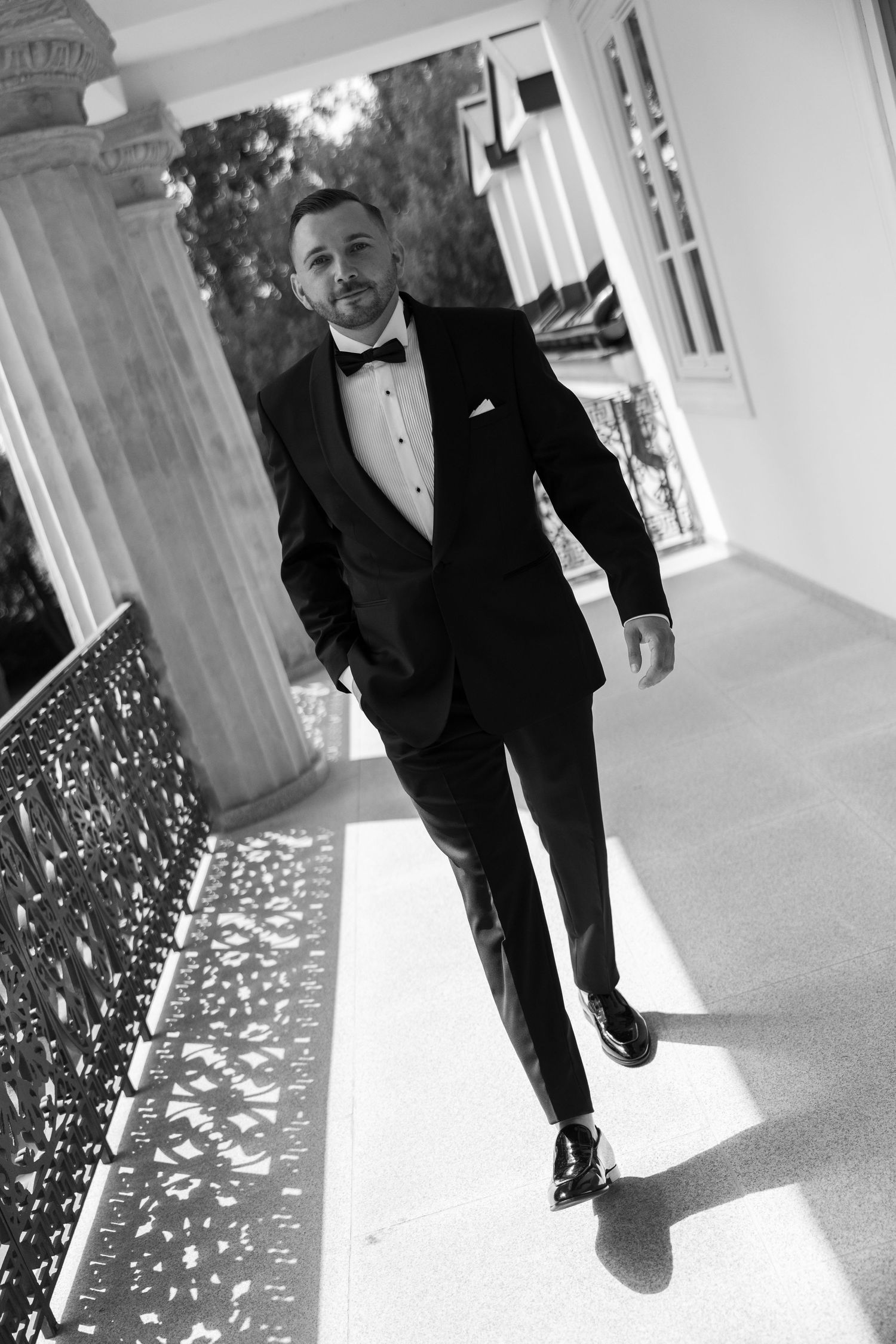 Black and white photo of a well-dressed man in a tuxedo with bow tie walking confidently along an elegant columned veranda.
