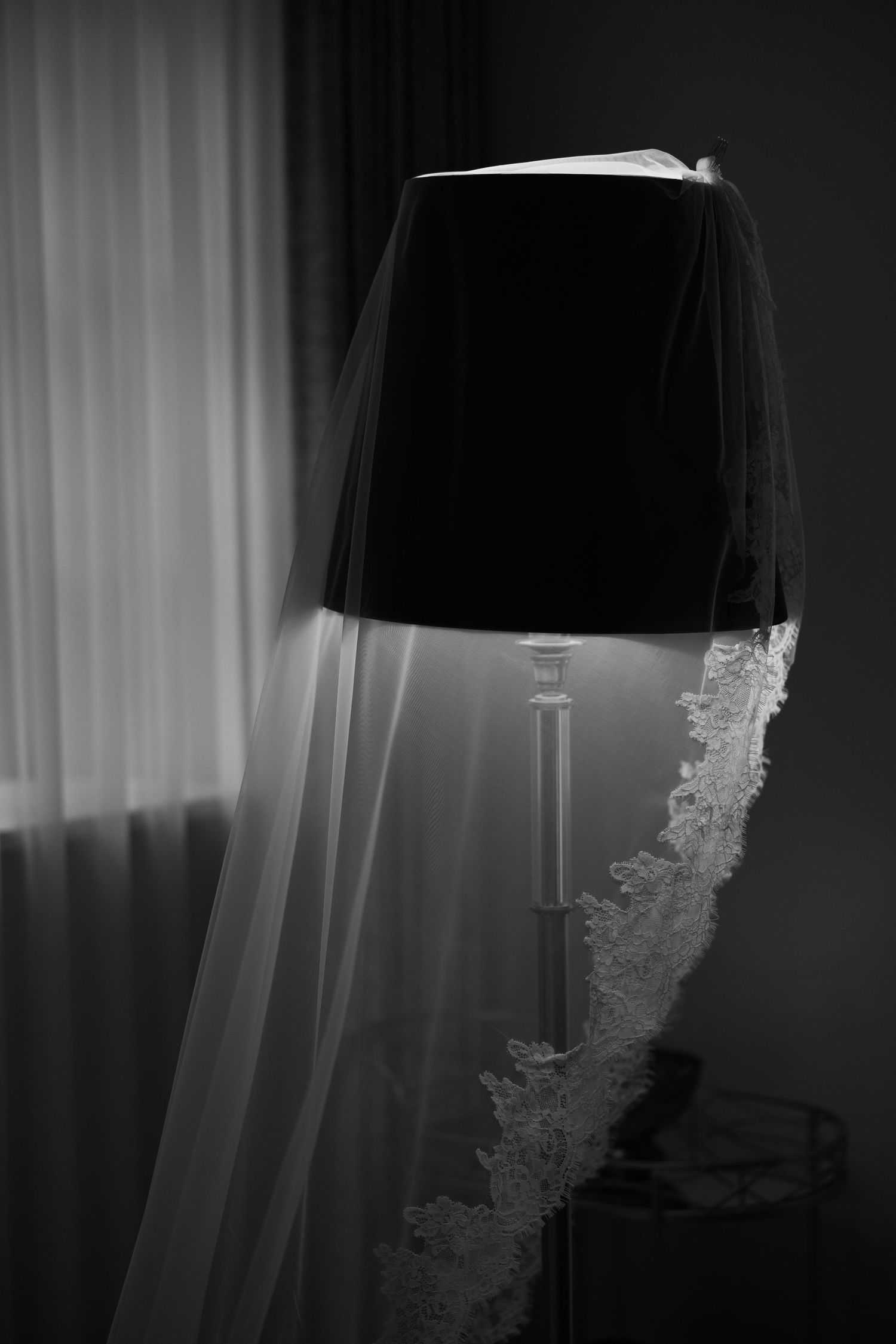 Black and white photo of a wedding veil draped over a lamp with lace trim detail near a window.
