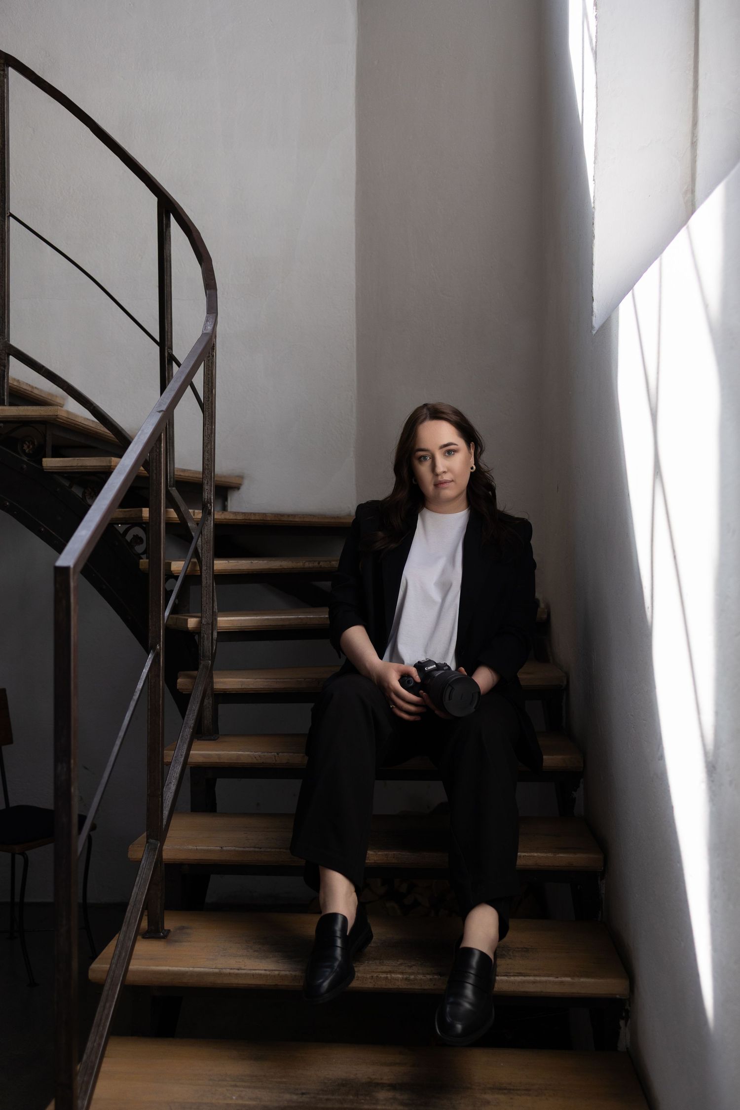 A young woman in a black blazer sits on wooden stairs holding a camera, looking at the camera in a modern interior.