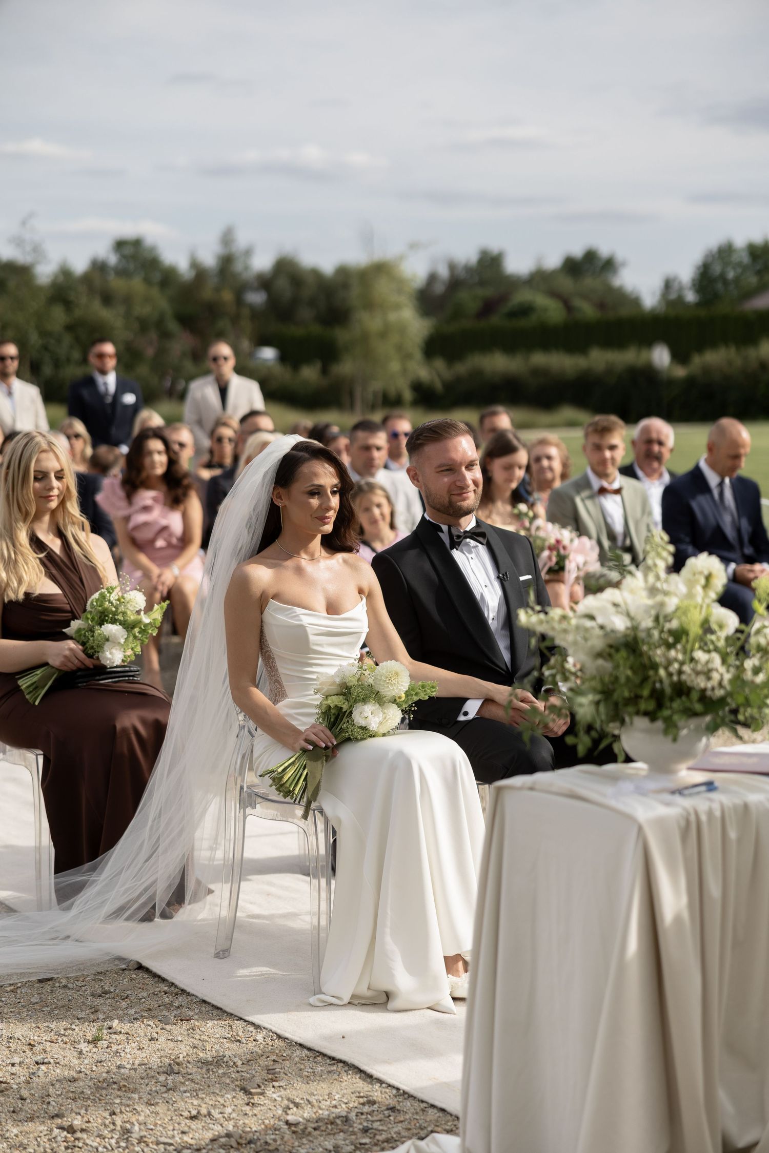 Bride and groom seated at outdoor wedding ceremony with guests, bride in white gown holding bouquet.