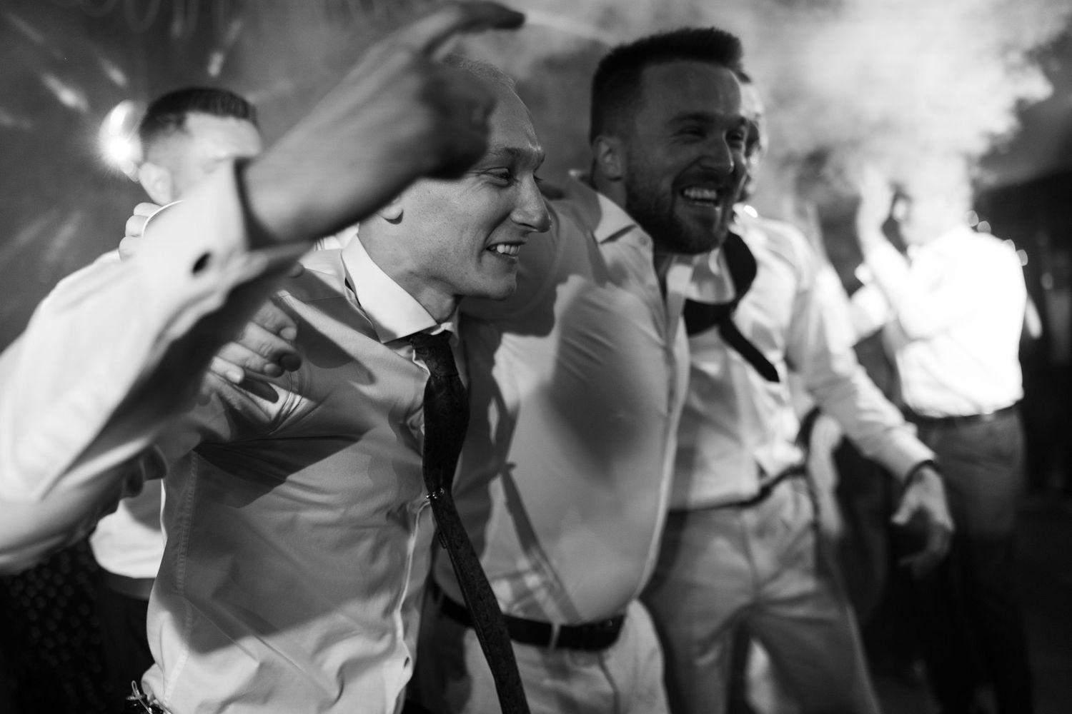 Men in dress shirts and suspenders dancing joyfully at a wedding reception in black and white photo.