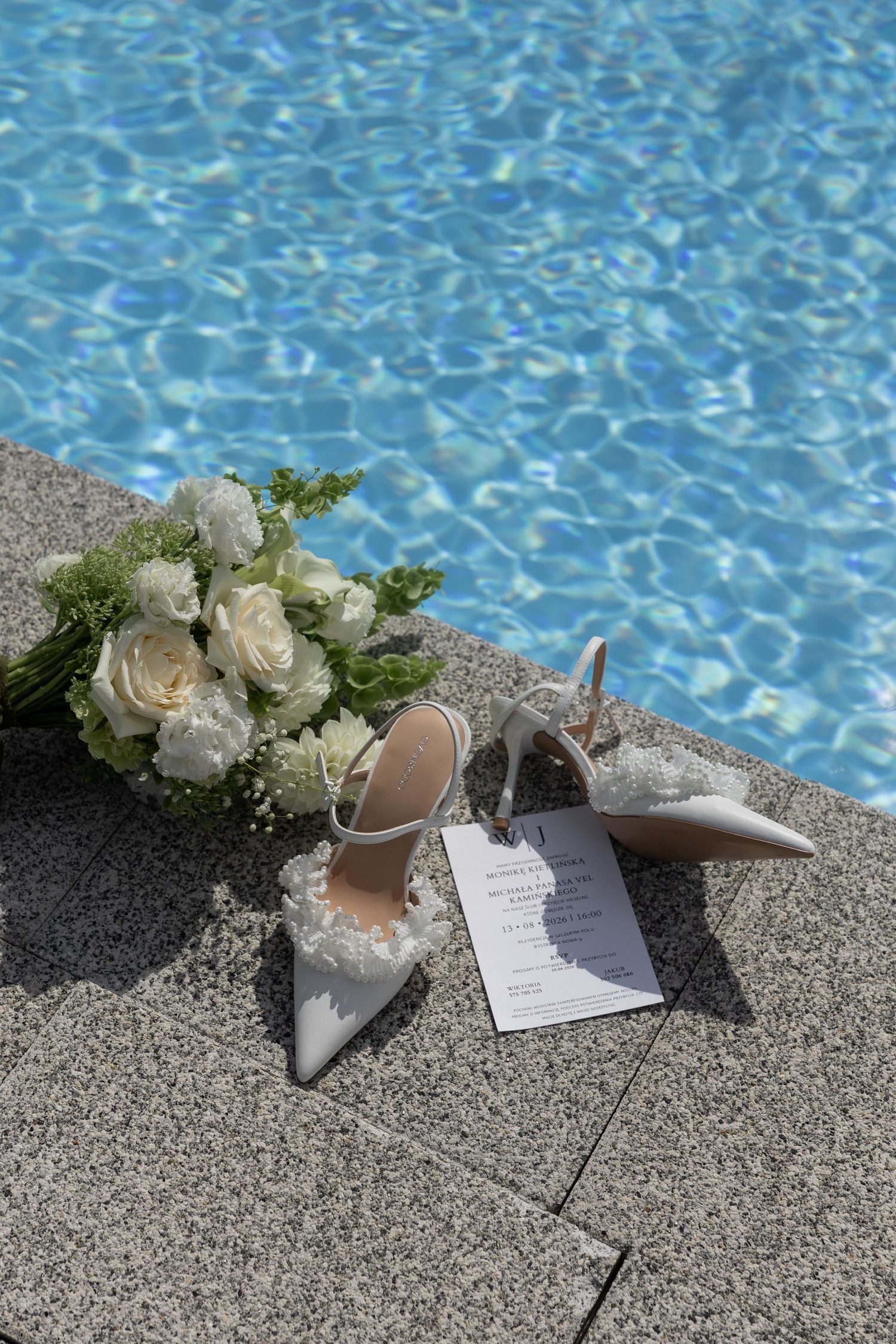 White bridal heels and floral bouquet with roses beside a sparkling blue swimming pool on a sunny day.