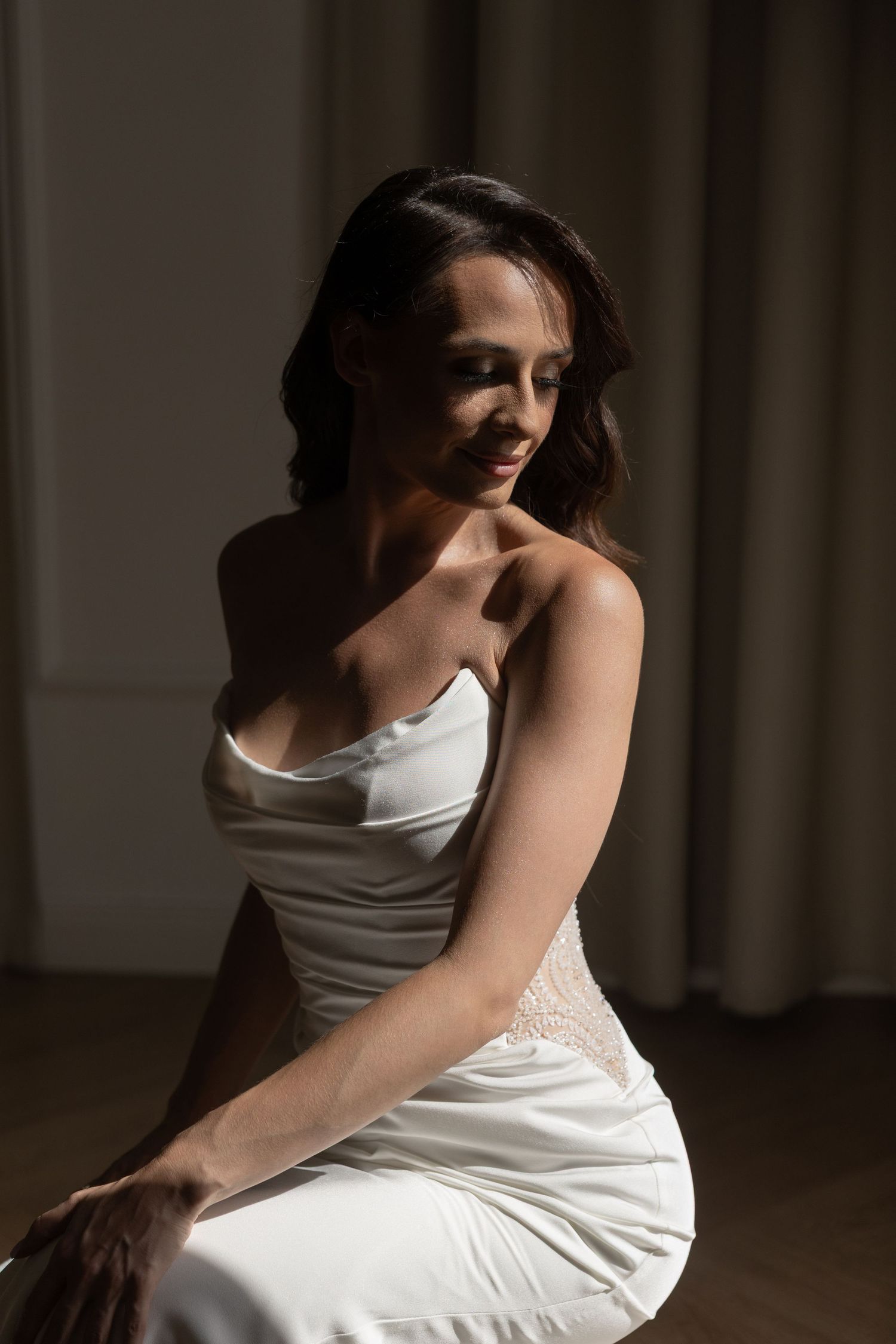 Elegant woman in white satin strapless gown sitting gracefully in soft dramatic lighting.