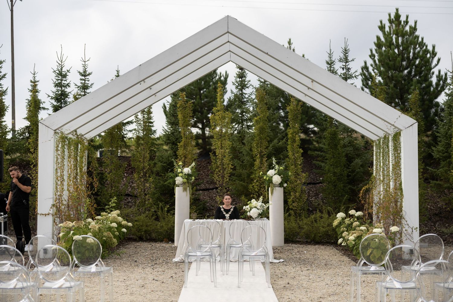 Elegant outdoor wedding ceremony setup with white geometric arch, floral arrangements, and ghost chairs amid pine trees.