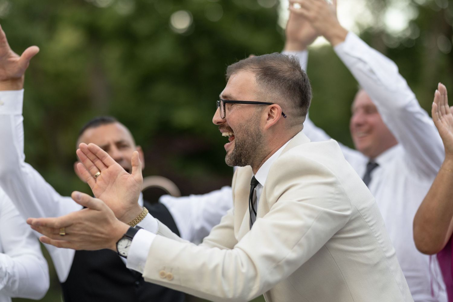 Man in white suit clapping and laughing at outdoor wedding celebration with guests in background.