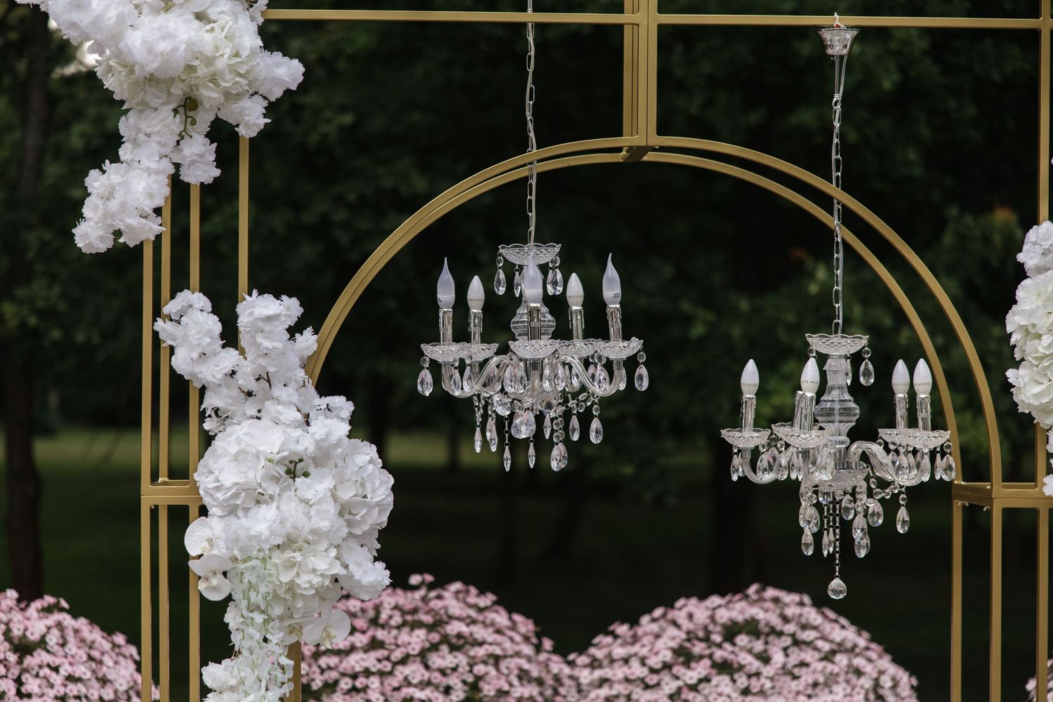 Crystal chandeliers hang from a gold arch wedding backdrop adorned with white and pink floral arrangements outdoors.
