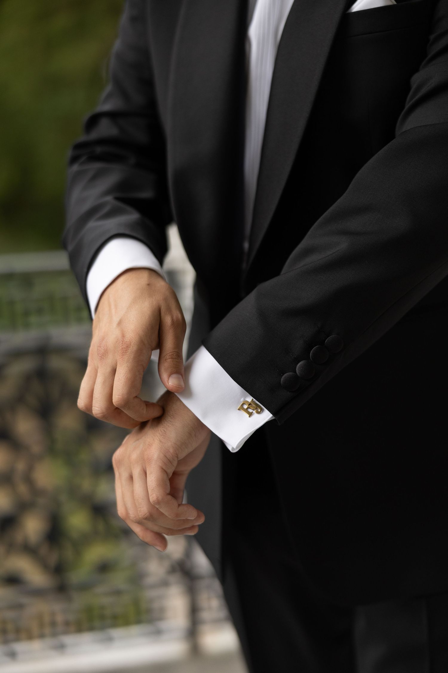Man in black tuxedo adjusting gold cufflinks on white dress shirt sleeve outdoors.