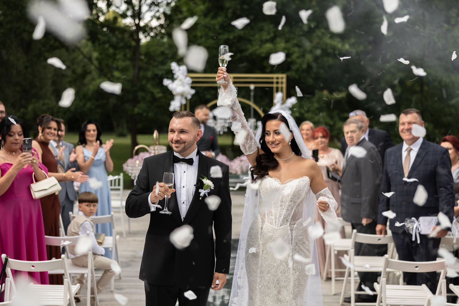Joyful newlyweds walk down the aisle as guests toss white confetti at an elegant outdoor wedding ceremony.