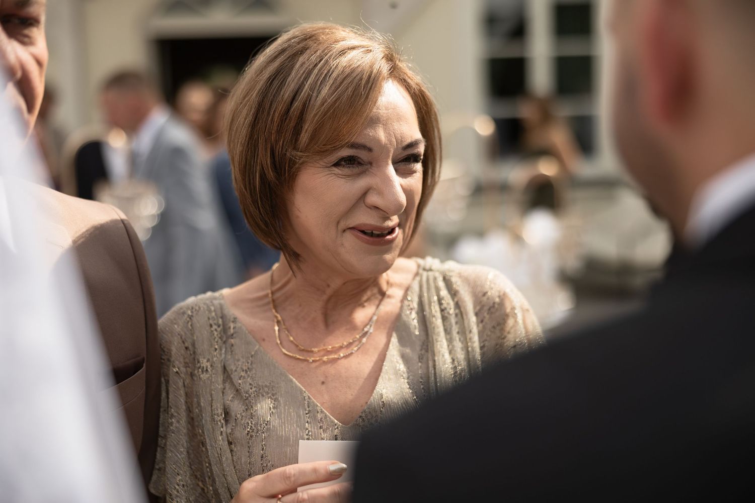 Elegant older woman with bob haircut wearing gold necklace and beige lace top, socializing at a formal outdoor event.