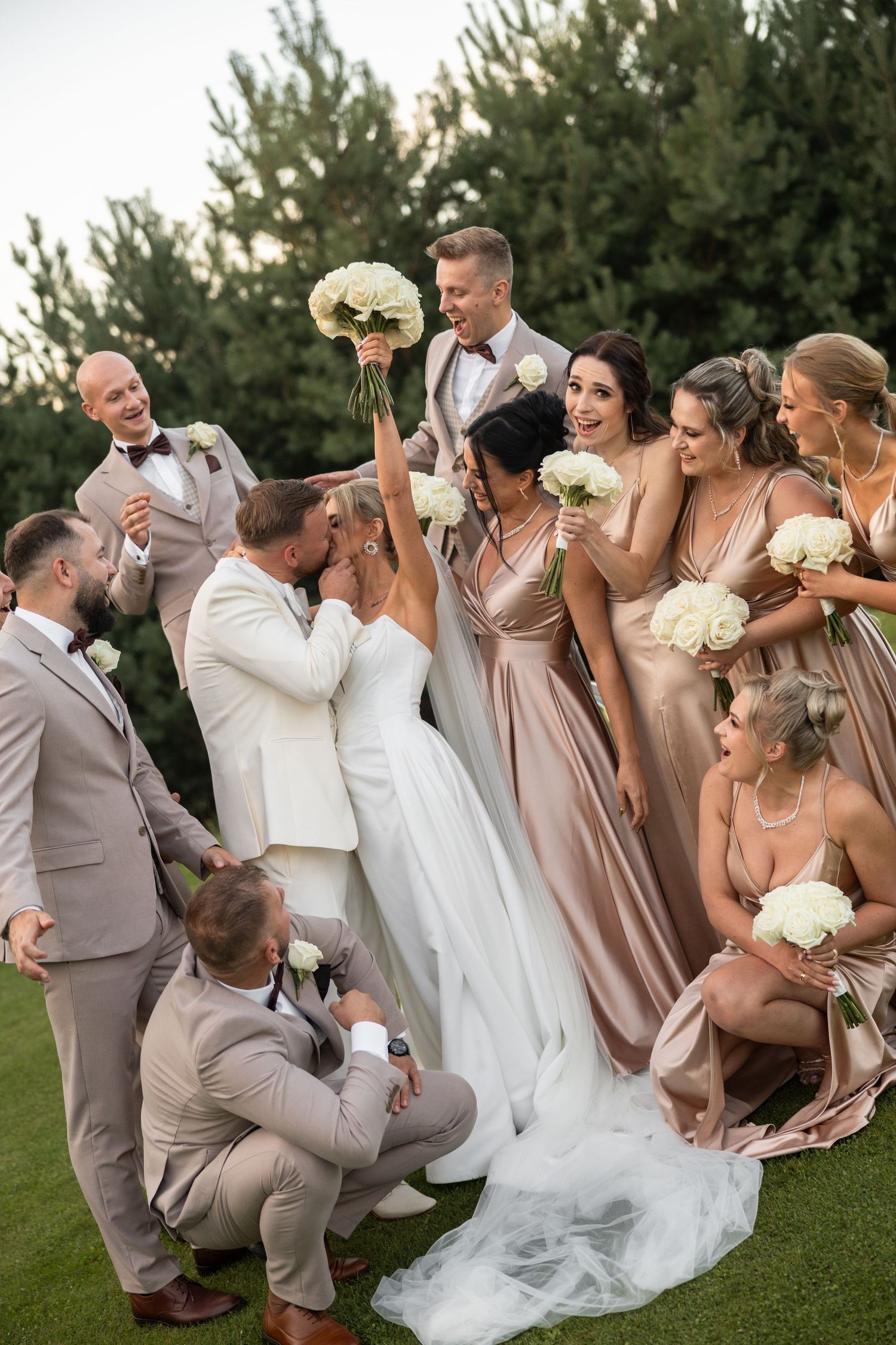 Wedding party celebrates as bride and groom kiss, surrounded by bridesmaids in blush gowns and groomsmen on a green lawn.