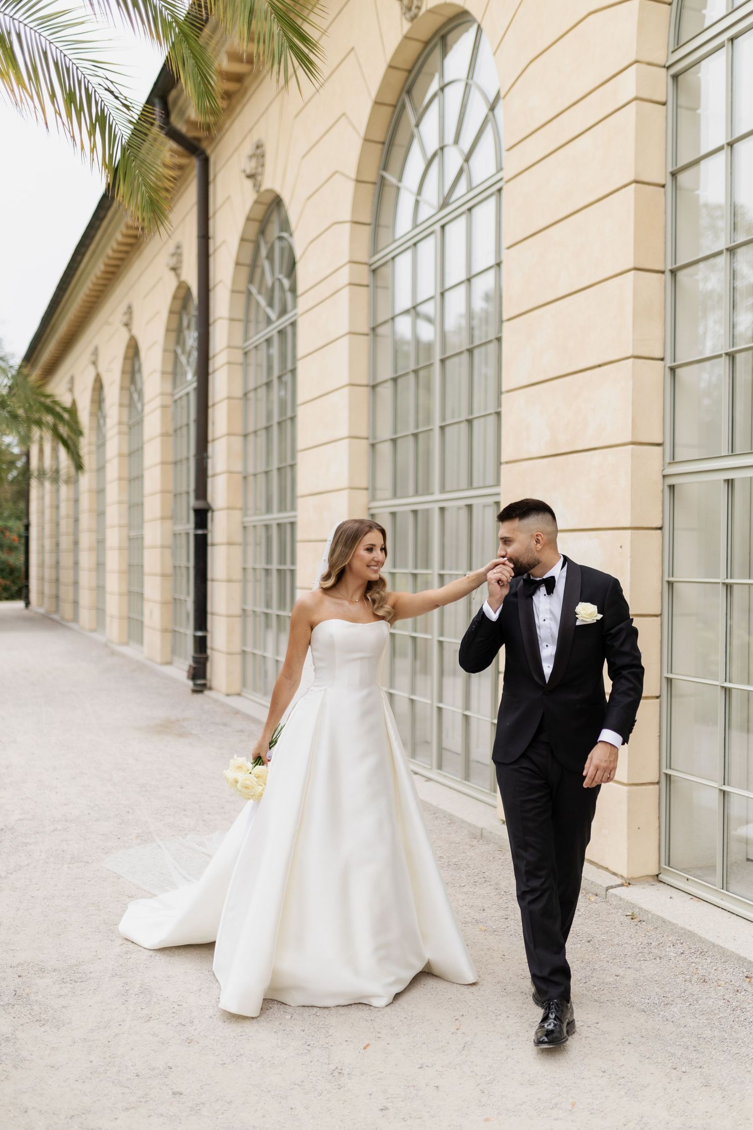 Elegant couple walking along arched building exterior with bride in classic white gown.