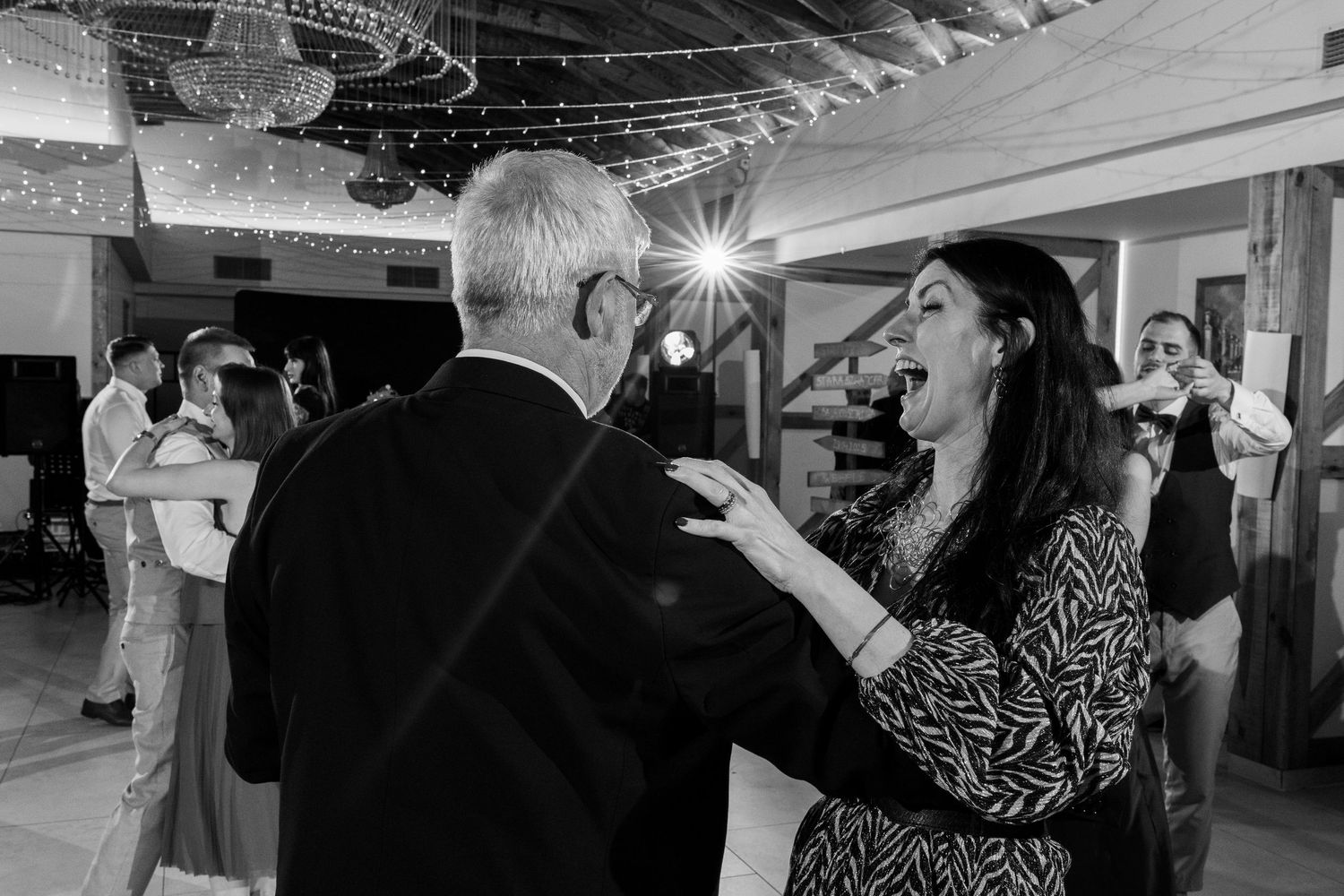 A couple dances together at a party while others mingle in the background of a dimly lit room.