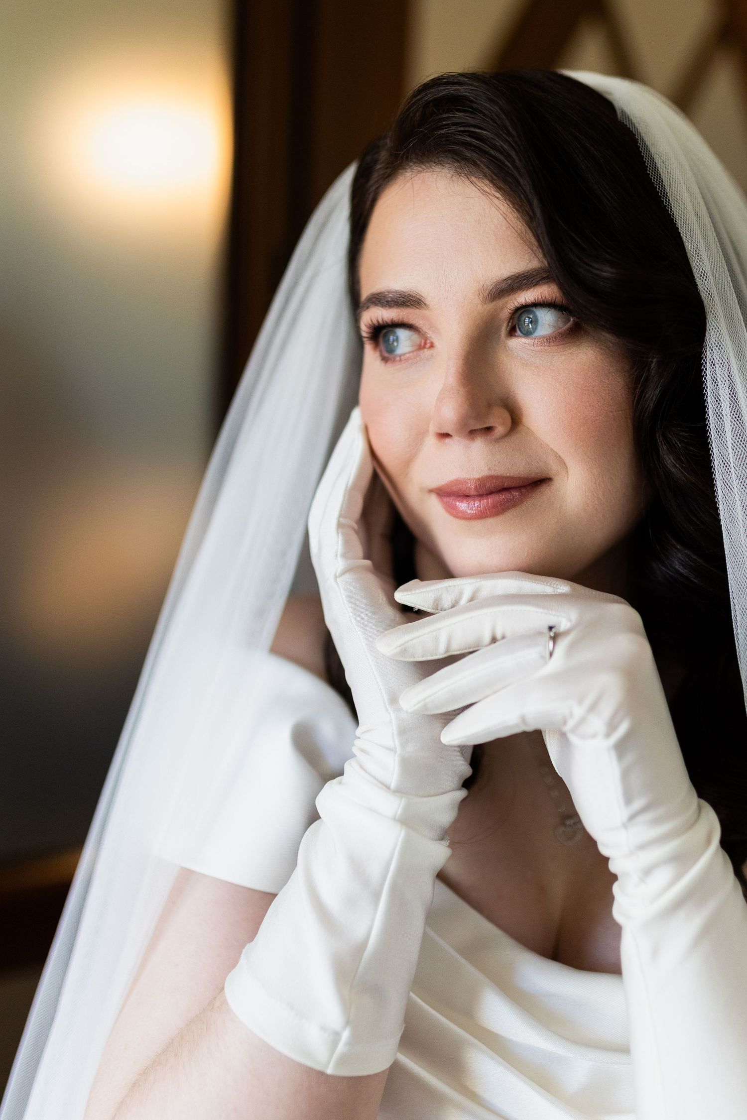 A bride in white gloves and veil poses elegantly with a gentle smile in beautiful bridal portrait lighting.