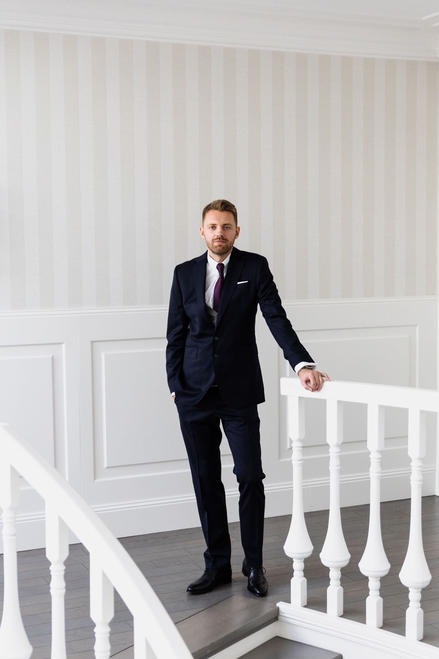 A person wearing a dark navy suit and red tie stands elegantly on white stairs with classic balustrades in a bright interior.
