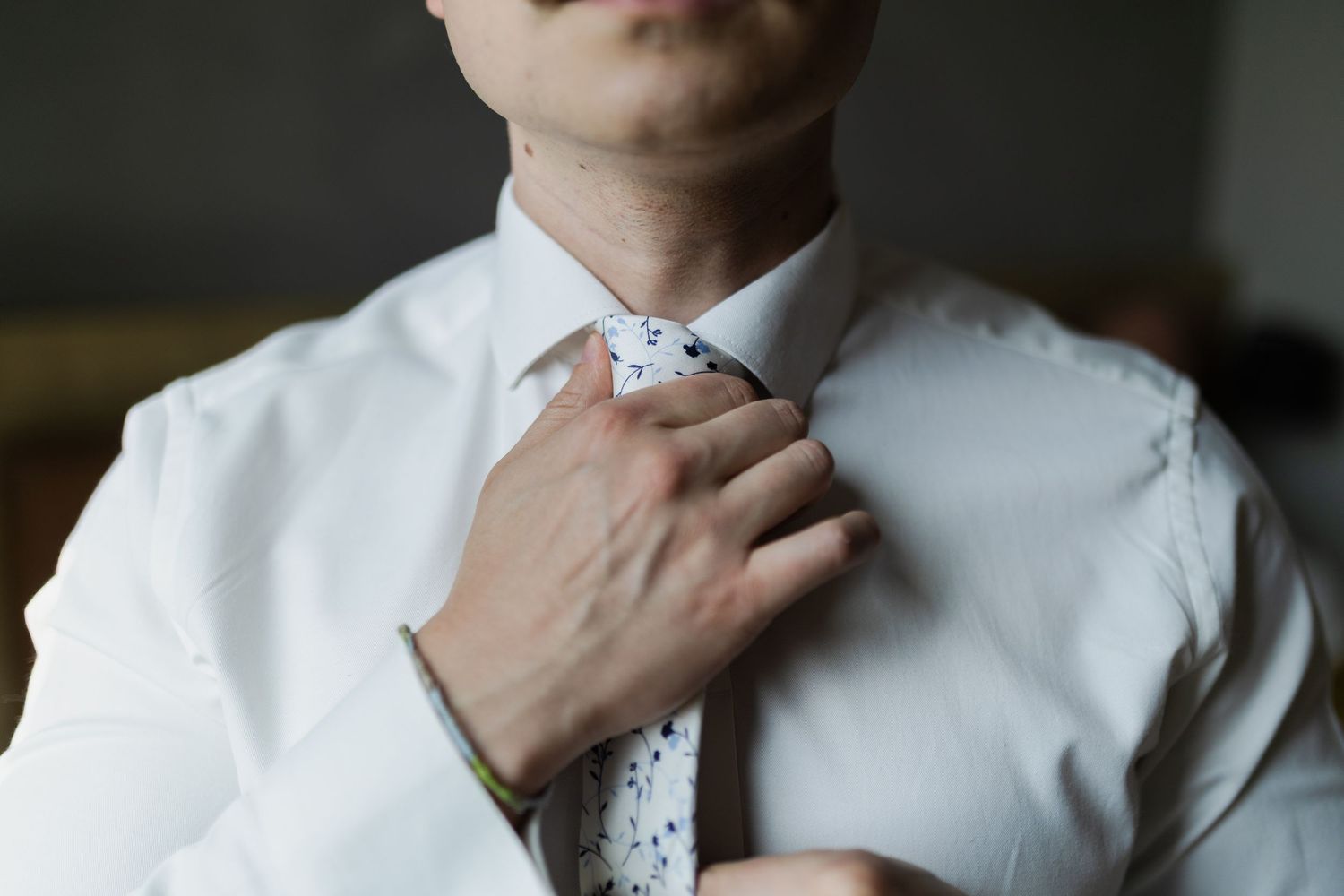 Close-up of hands adjusting a speckled necktie against crisp white dress shirt in dim lighting.