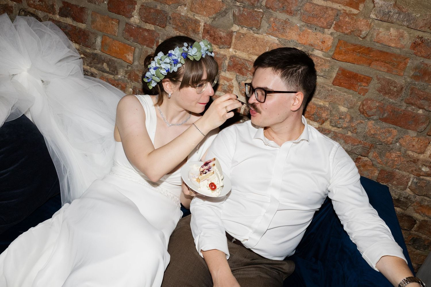 Newlyweds in casual wedding attire share a playful moment eating dessert against exposed brick wall.