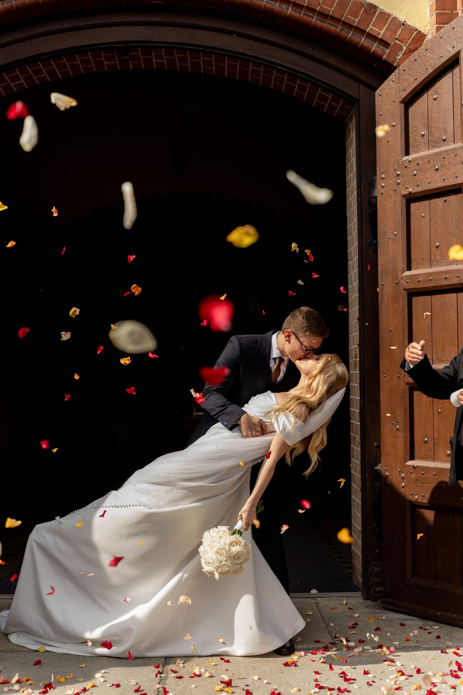 A romantic wedding couple dips in a dance pose as rose petals fall around them in front of wooden church doors.