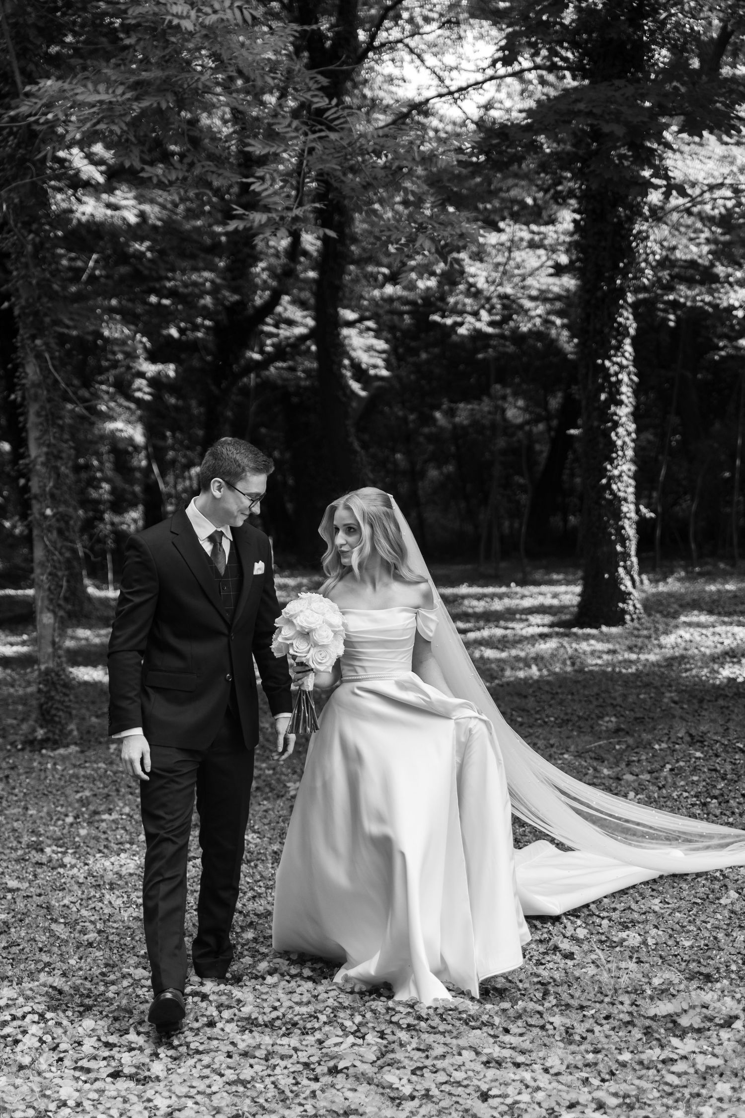A couple in wedding attire walks together on a path covered in fallen autumn leaves in a wooded setting.