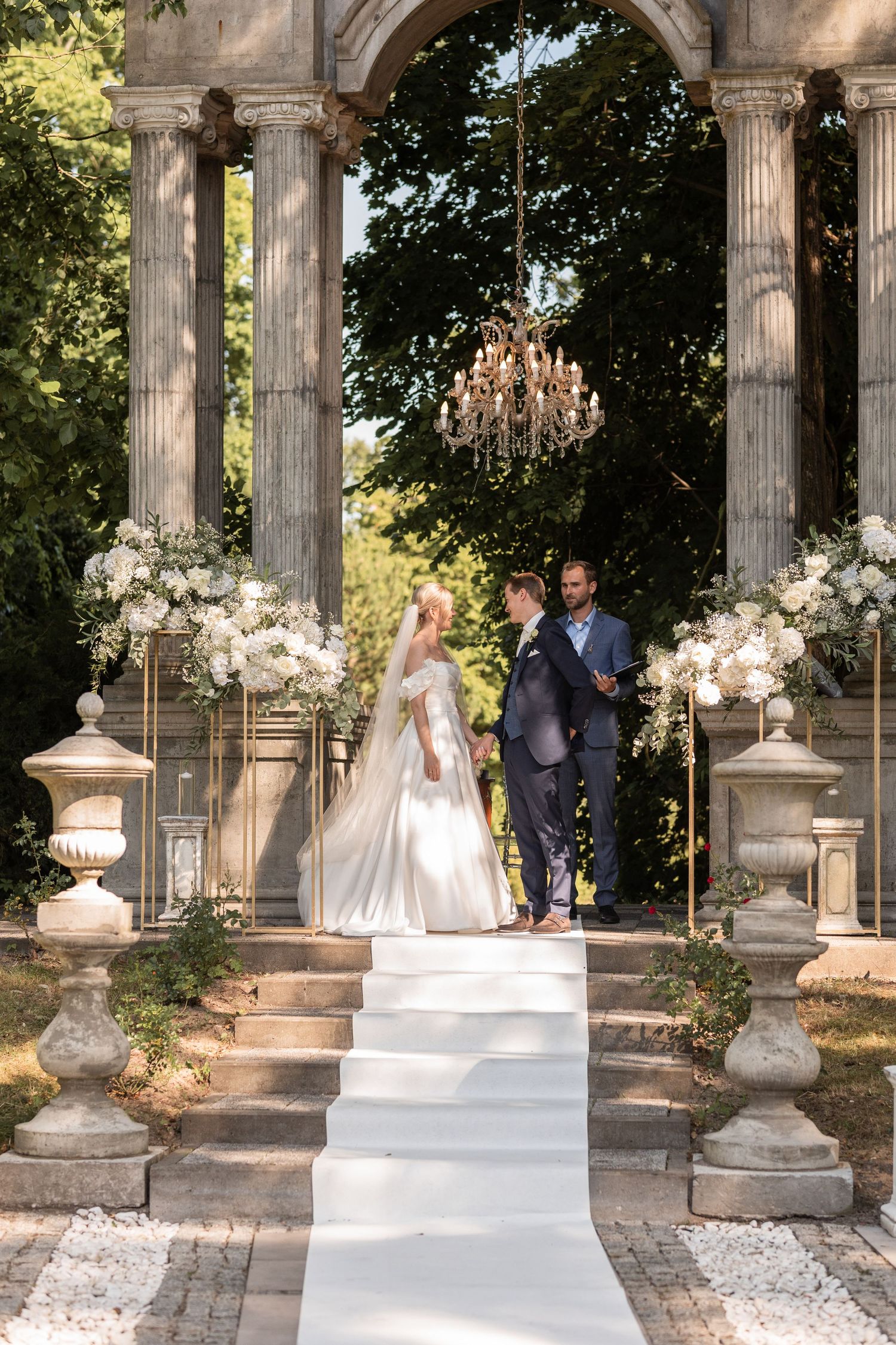 A wedding couple stands in an elegant outdoor garden setting with classical stone columns and floral arrangements.
