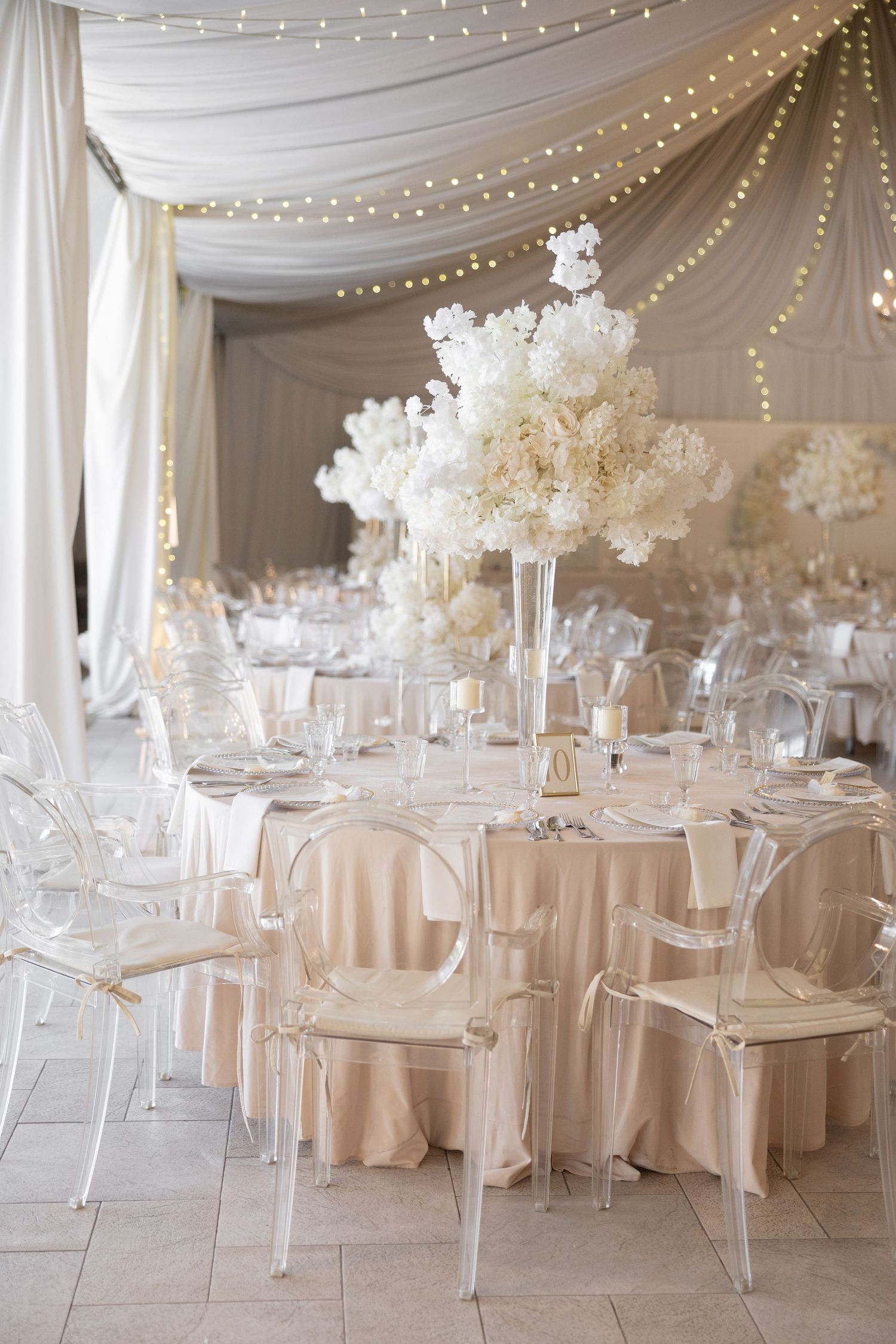 Elegant wedding reception with ghost chairs, blush linens, tall white floral centerpieces, and string lights under a draped tent.