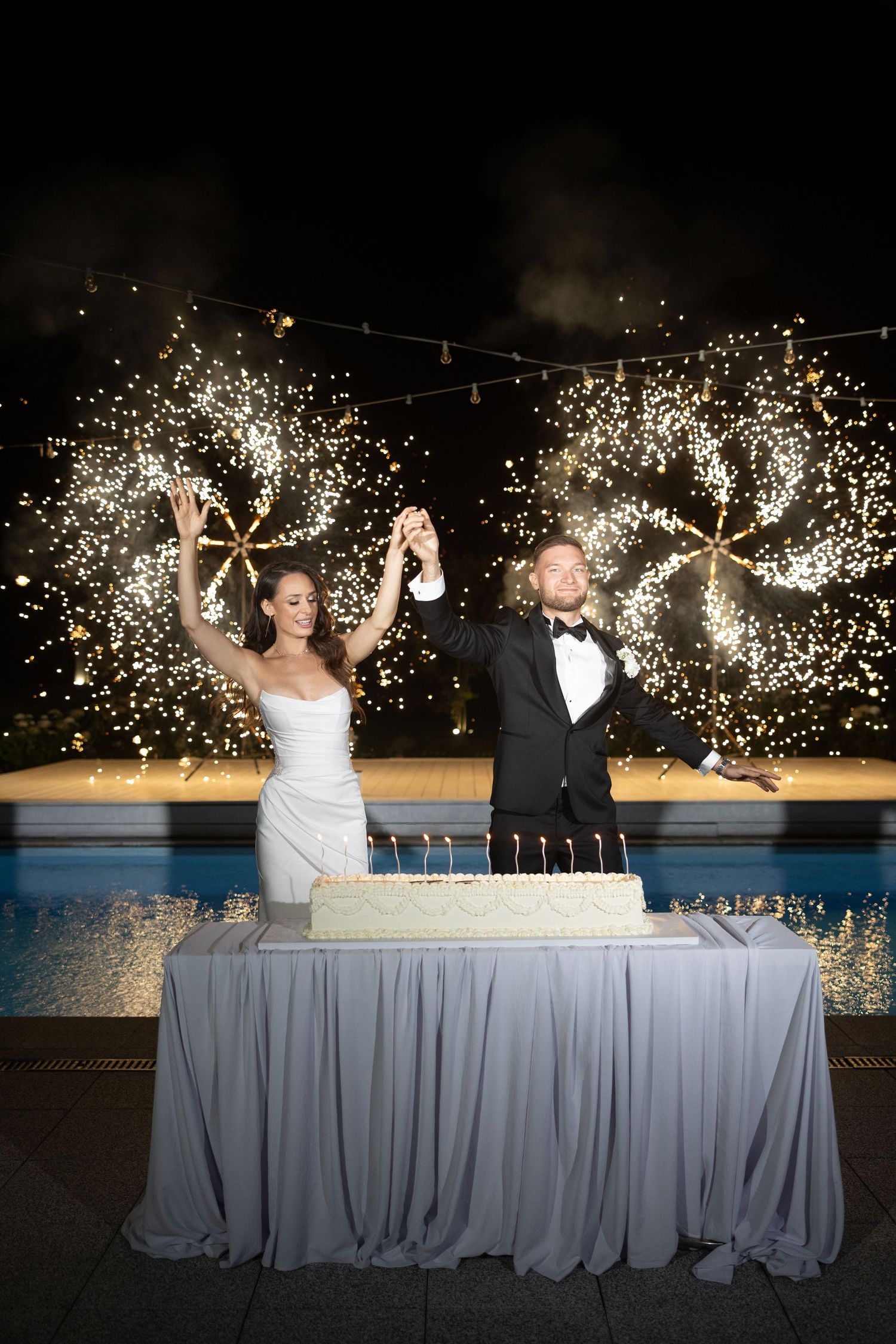 Bride and groom cutting wedding cake with sparklers and fireworks display behind them at night poolside celebration.