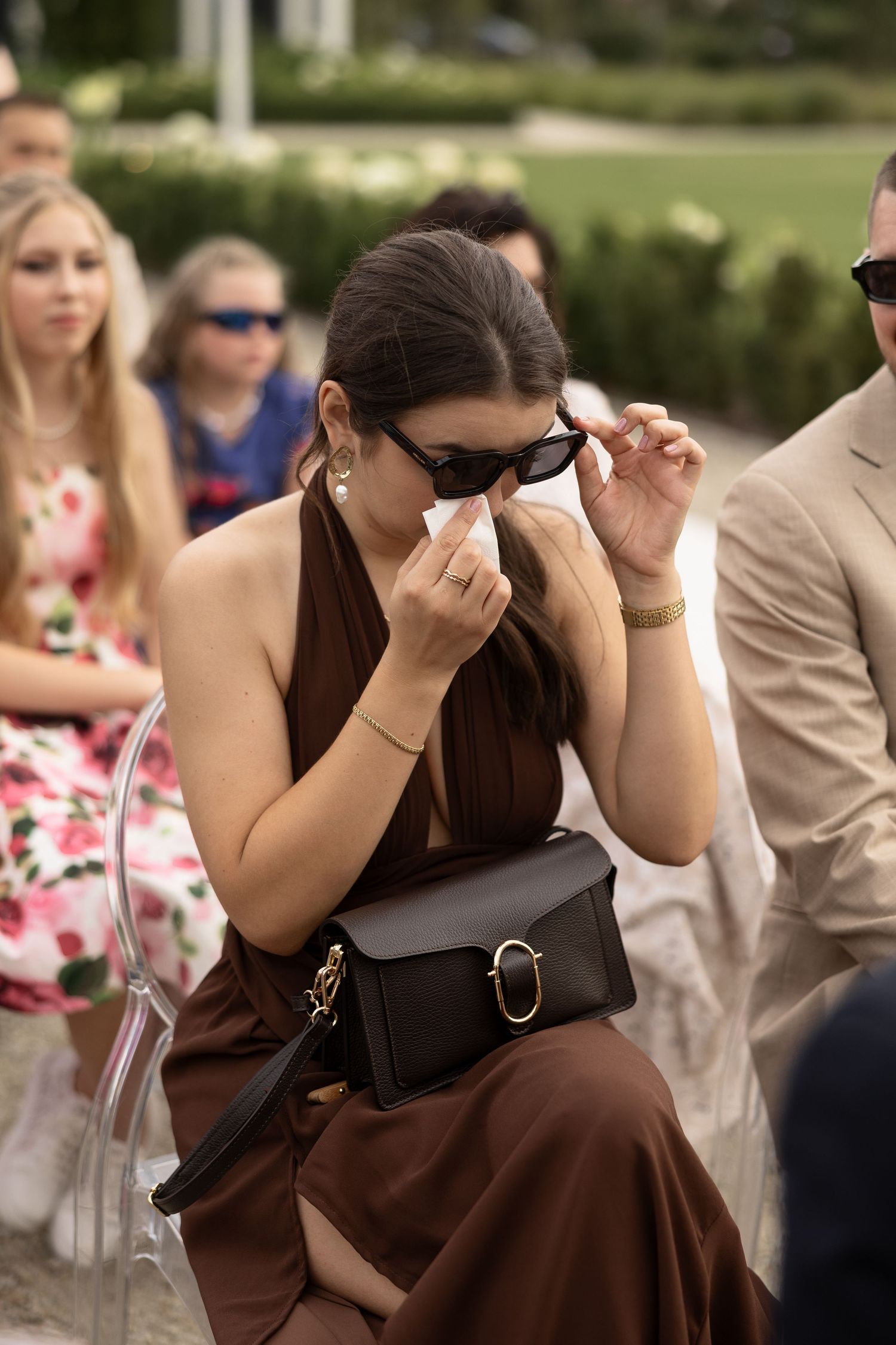 A woman in a brown dress wipes tears at an outdoor wedding ceremony, wearing sunglasses and holding a black crossbody bag.
