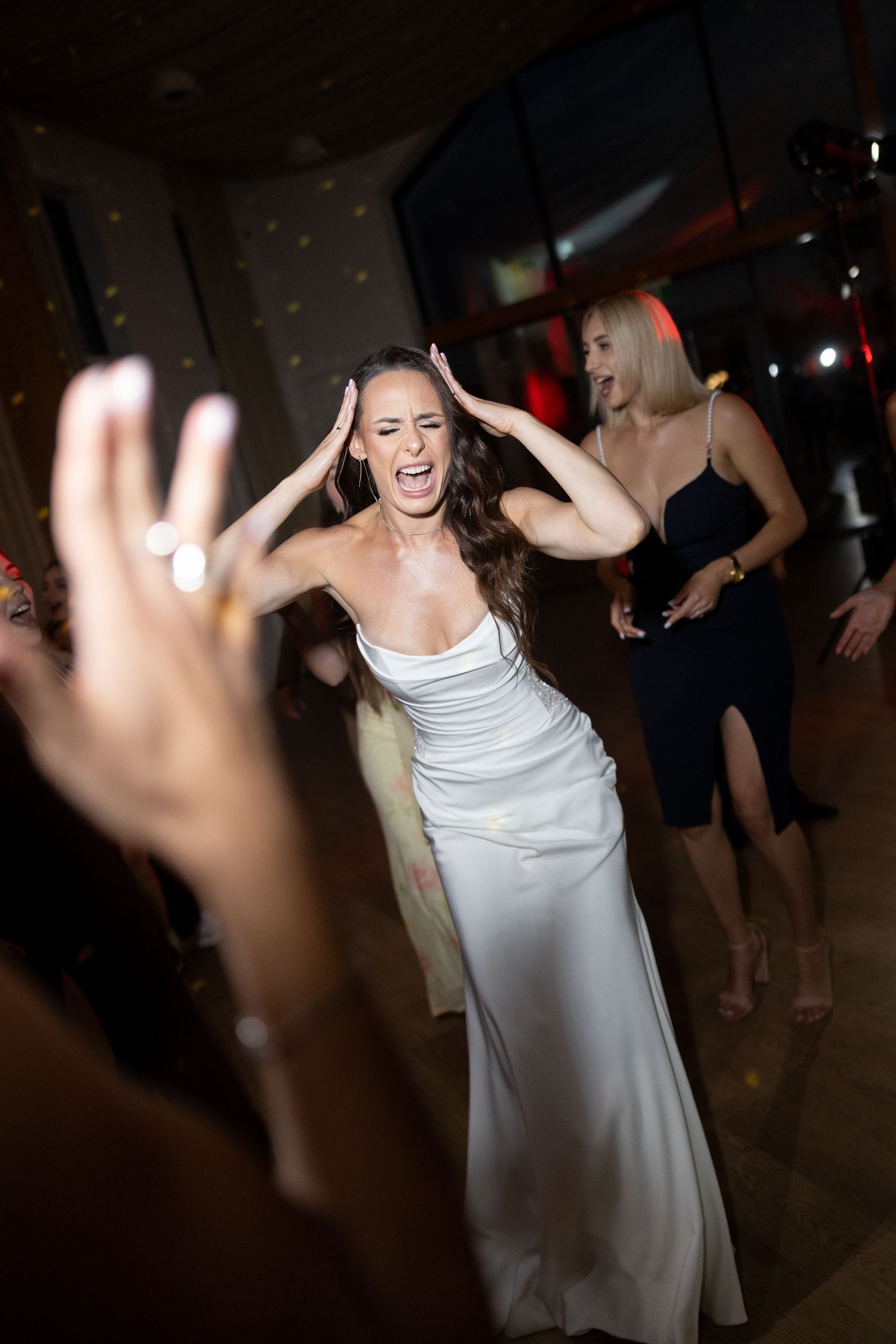 Bride in white gown dancing joyfully with hands on head at wedding reception, guests celebrating in background.