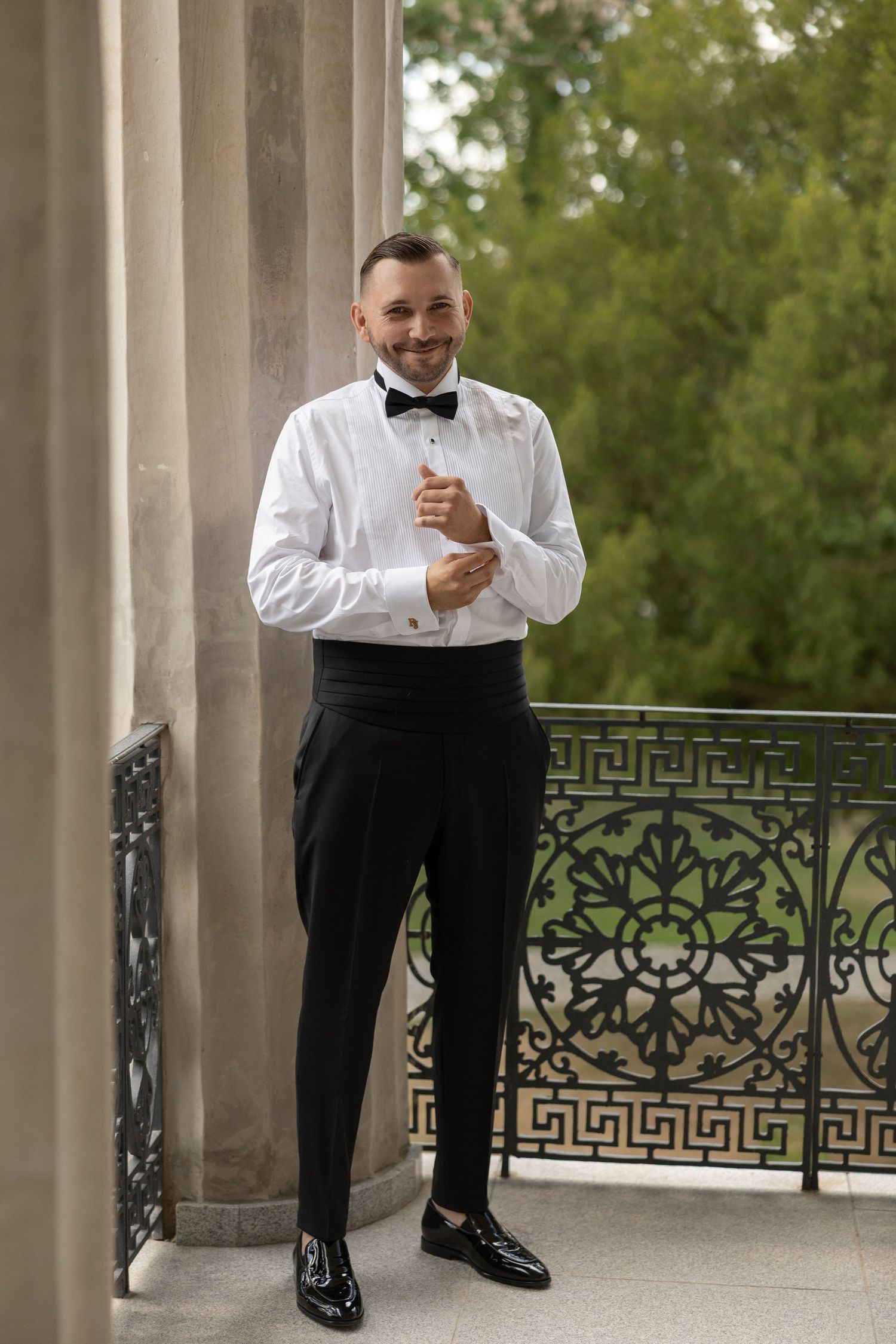 A smiling groom in a tuxedo adjusts his cufflinks on an ornate balcony surrounded by lush greenery.