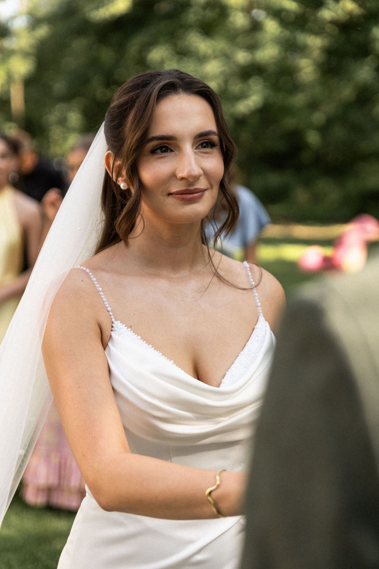A bride in an elegant white satin wedding dress with spaghetti straps and a flowing veil smiles in an outdoor garden setting.
