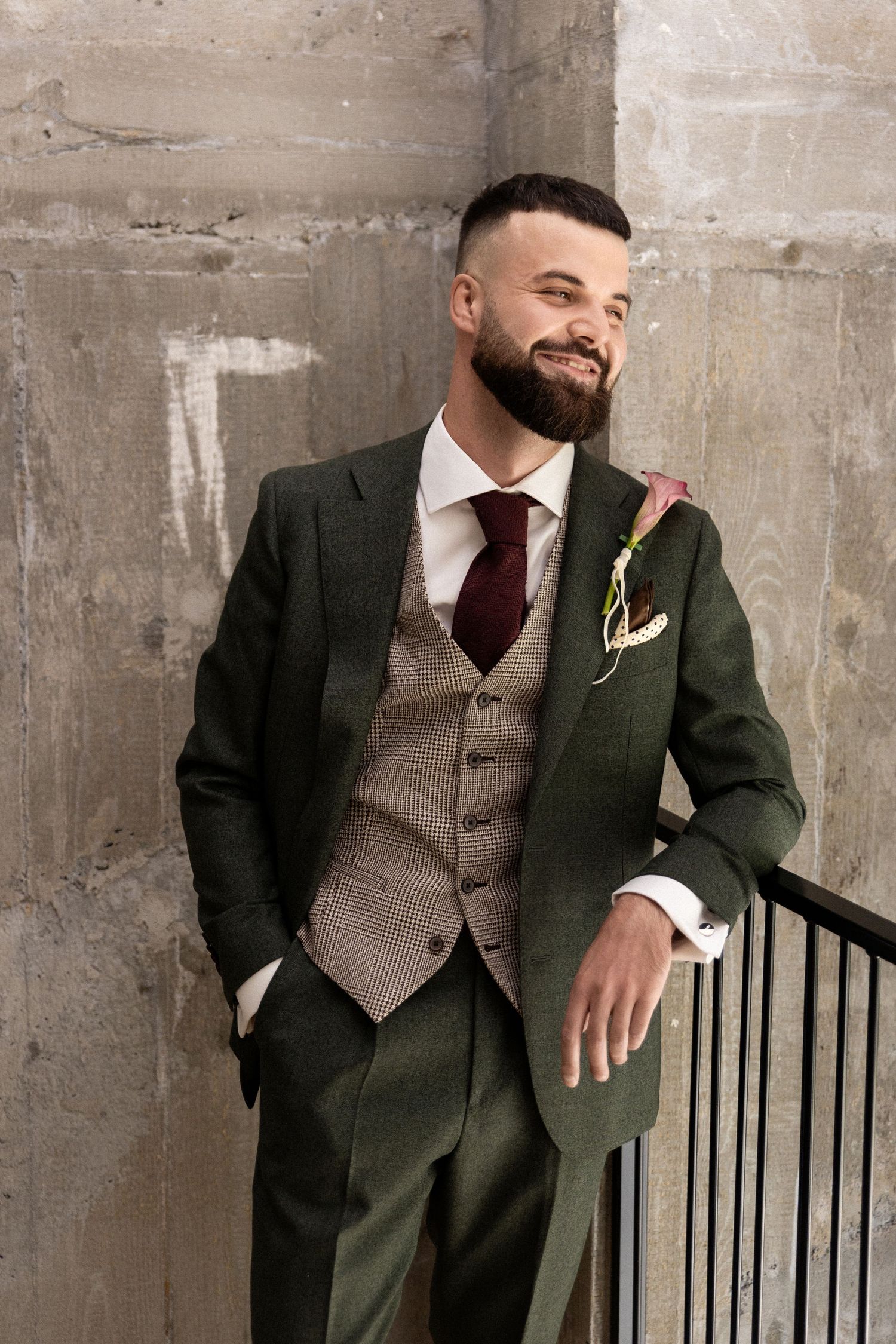 A groom in a dark green suit and brown vest poses against a concrete wall with a boutonniere.
