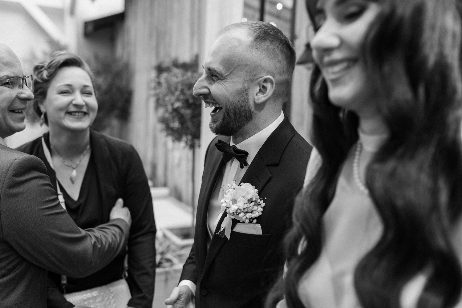 A candid black and white wedding moment captures joyful laughter shared between a groom and guests outdoors.