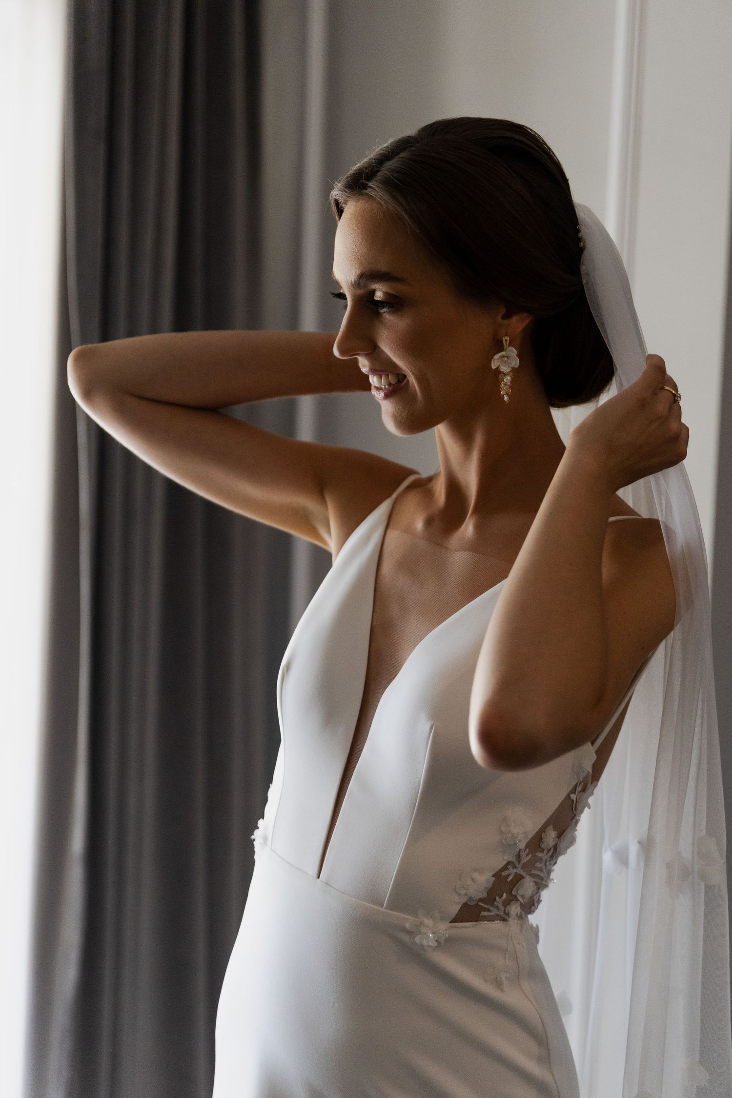 Bride wearing elegant sleeveless white wedding dress with plunging neckline and veil adjusts her hair by window.