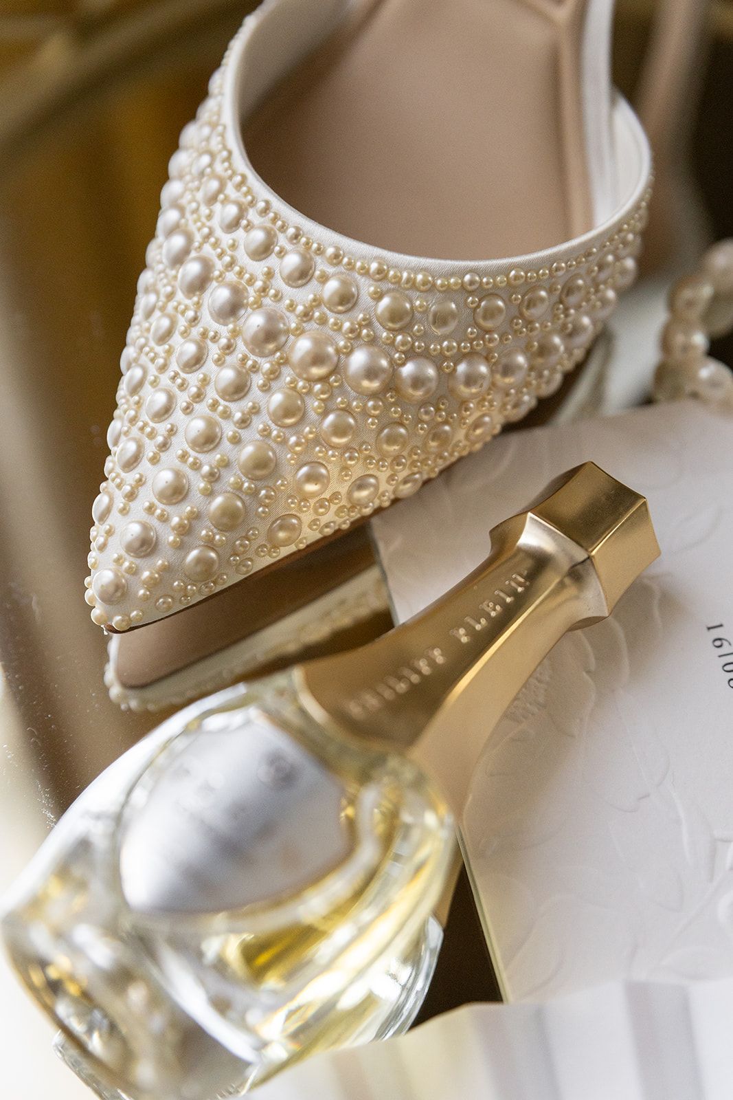 Elegant white bridal shoe decorated with pearls and crystals resting on perfume bottle in soft lighting.