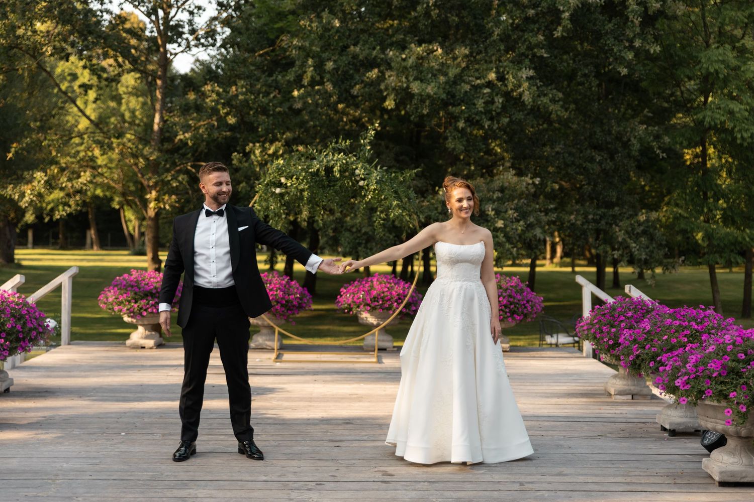 A wedding couple poses playfully in a park with vibrant pink flowers and lush greenery at sunset.