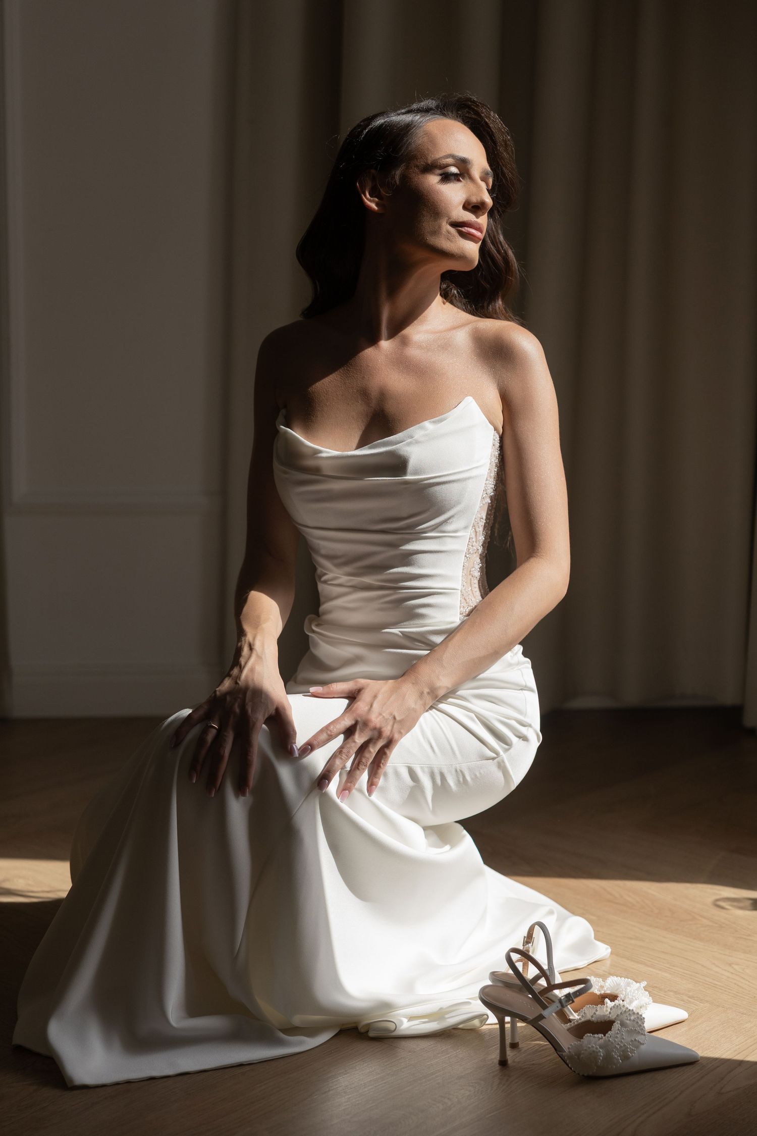 Elegant bride in a white strapless satin gown kneeling on wooden floor with bridal heels nearby in soft natural light.