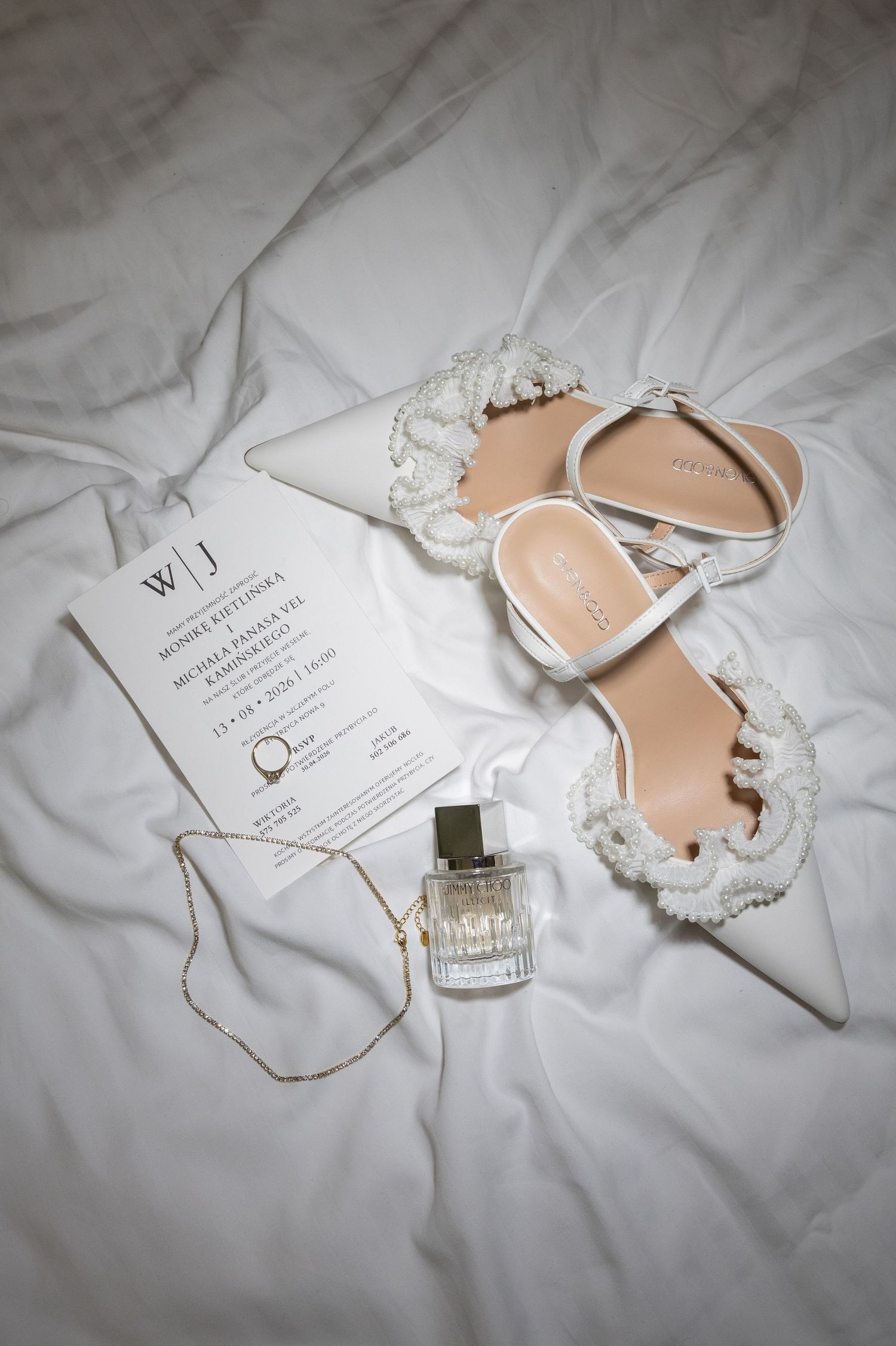 White pearl-adorned bridal heels, wedding invitation, gold necklace, and perfume bottle arranged on white satin fabric.