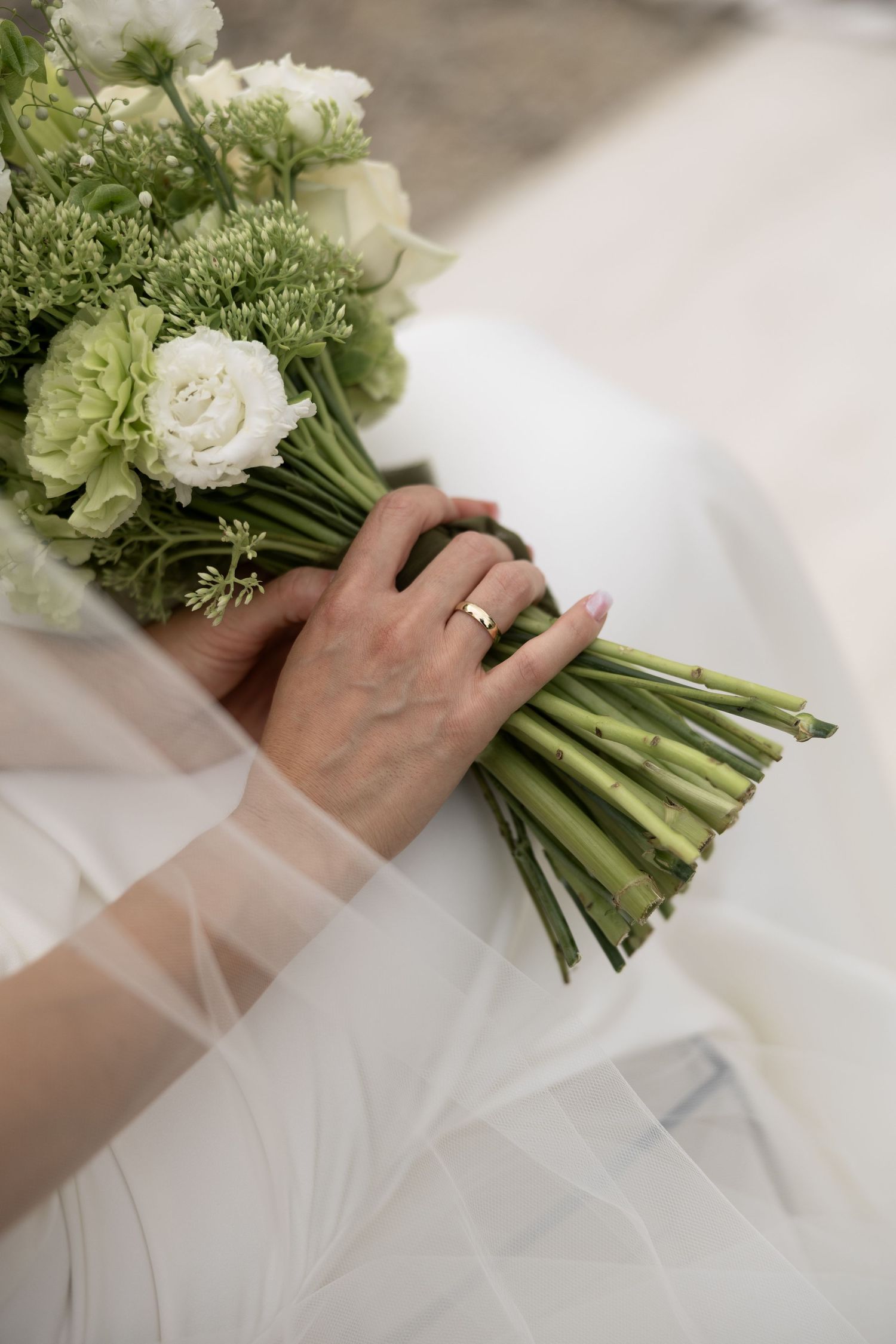 Bride holding a white and green wedding bouquet with a gold ring on her finger, draped in a flowing white veil.