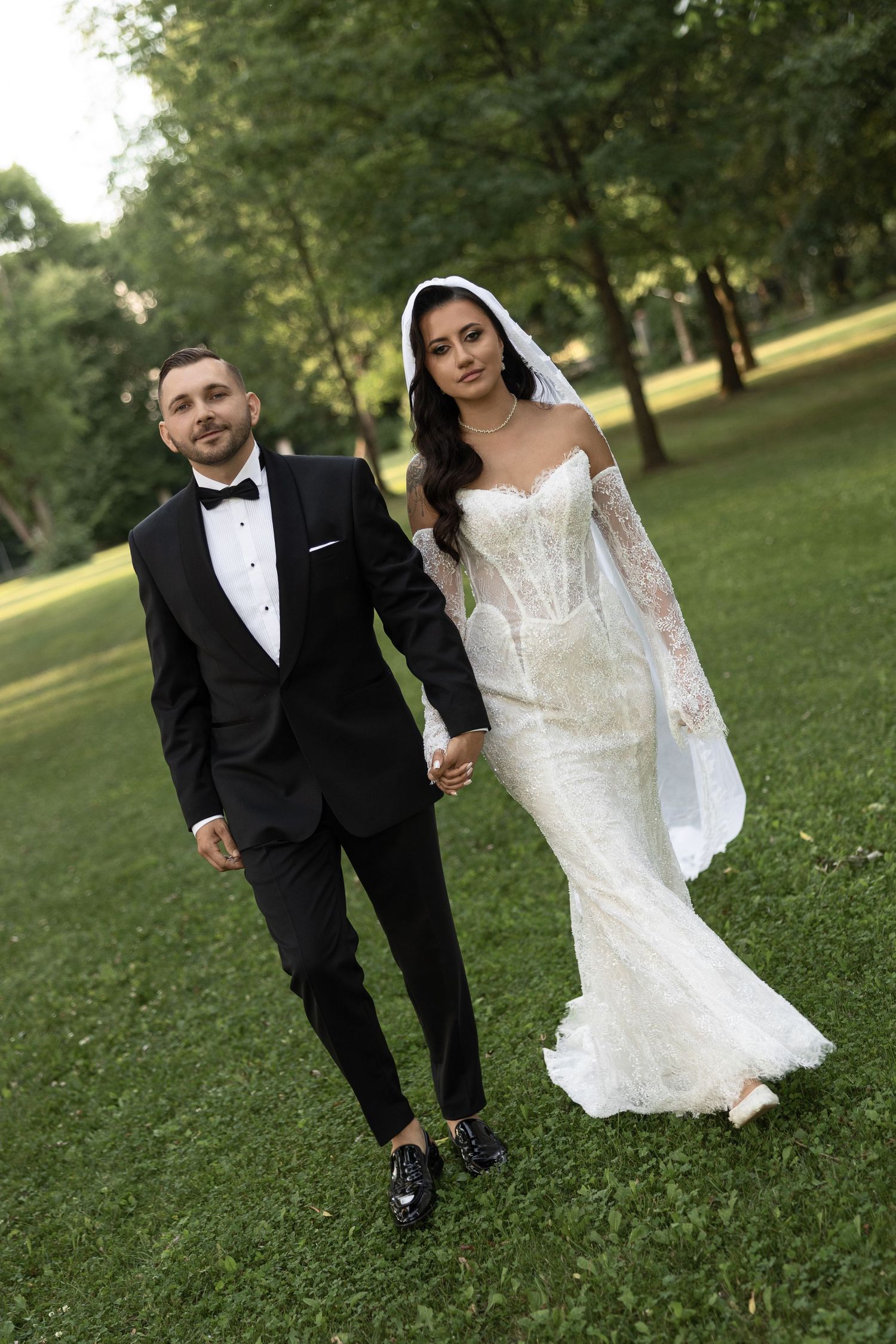Bride in white lace gown and veil walks hand-in-hand with groom in black tuxedo through lush green park.