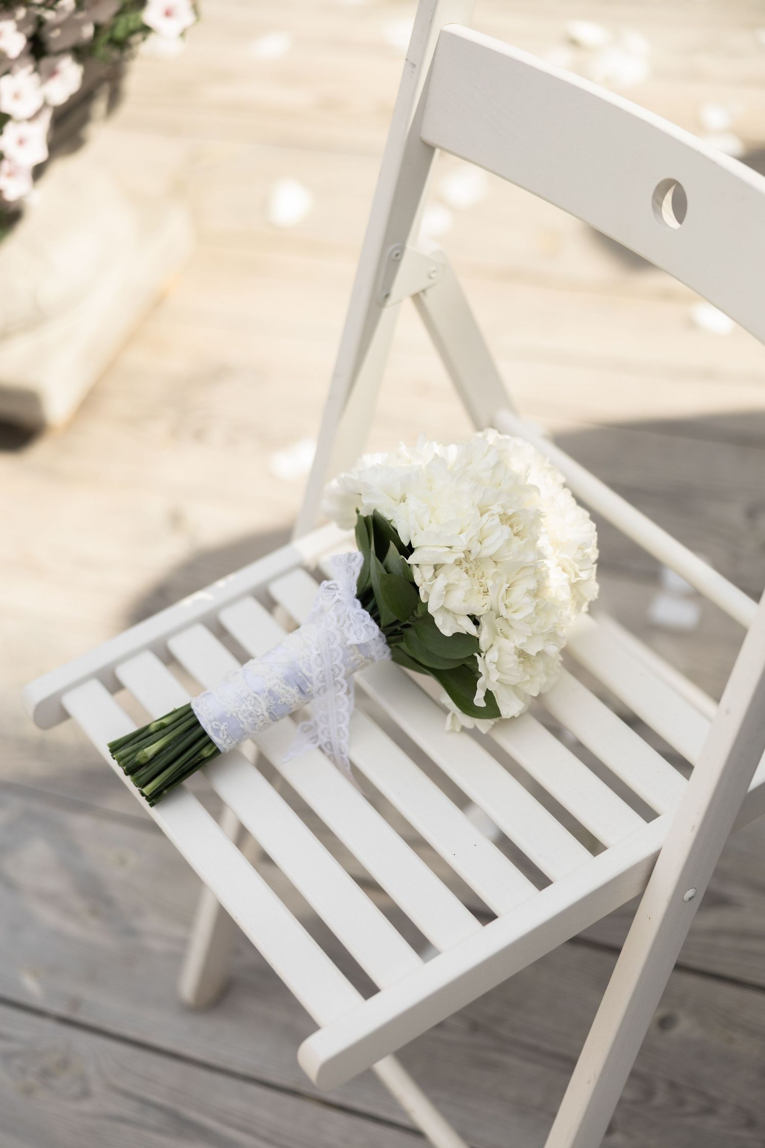 White hydrangea bridal bouquet with lace ribbon resting on a white folding chair at an outdoor wedding ceremony.
