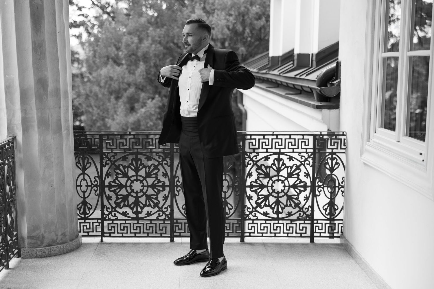 A man in a black tuxedo adjusts his bow tie on an ornate balcony in a classic black and white photo.