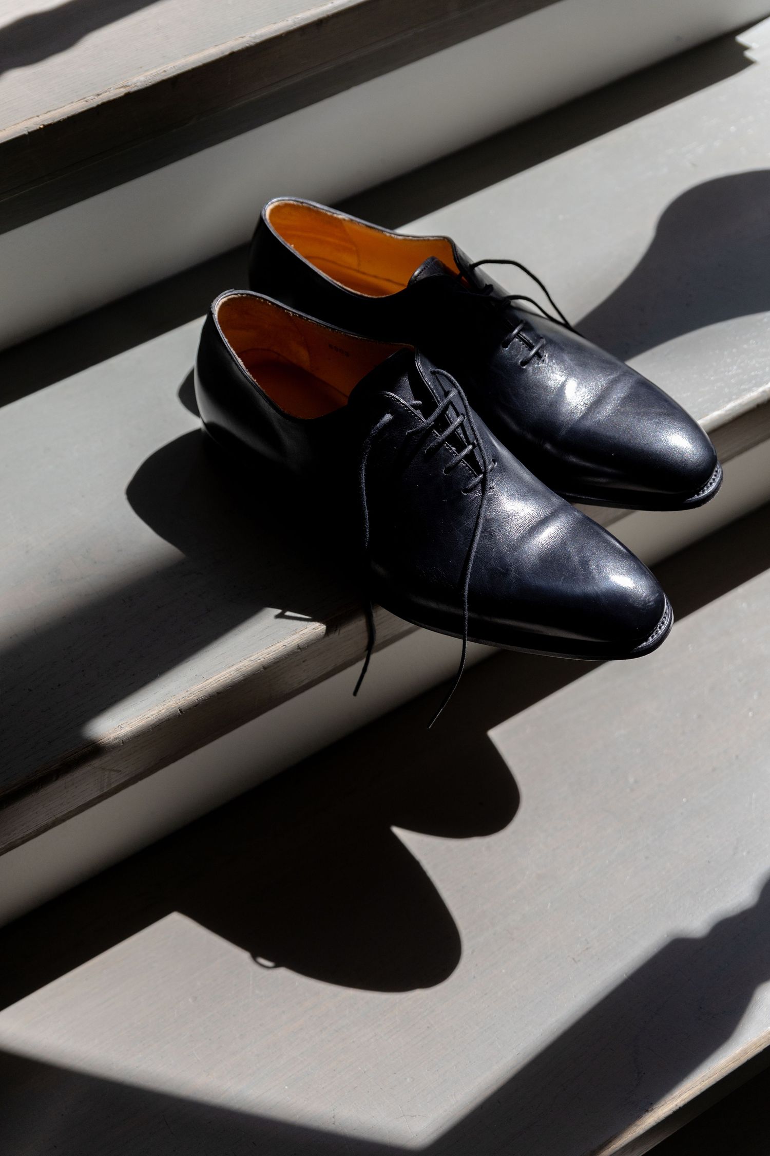 A pair of black leather oxford dress shoes casting dramatic shadows on a white striped surface in natural light.