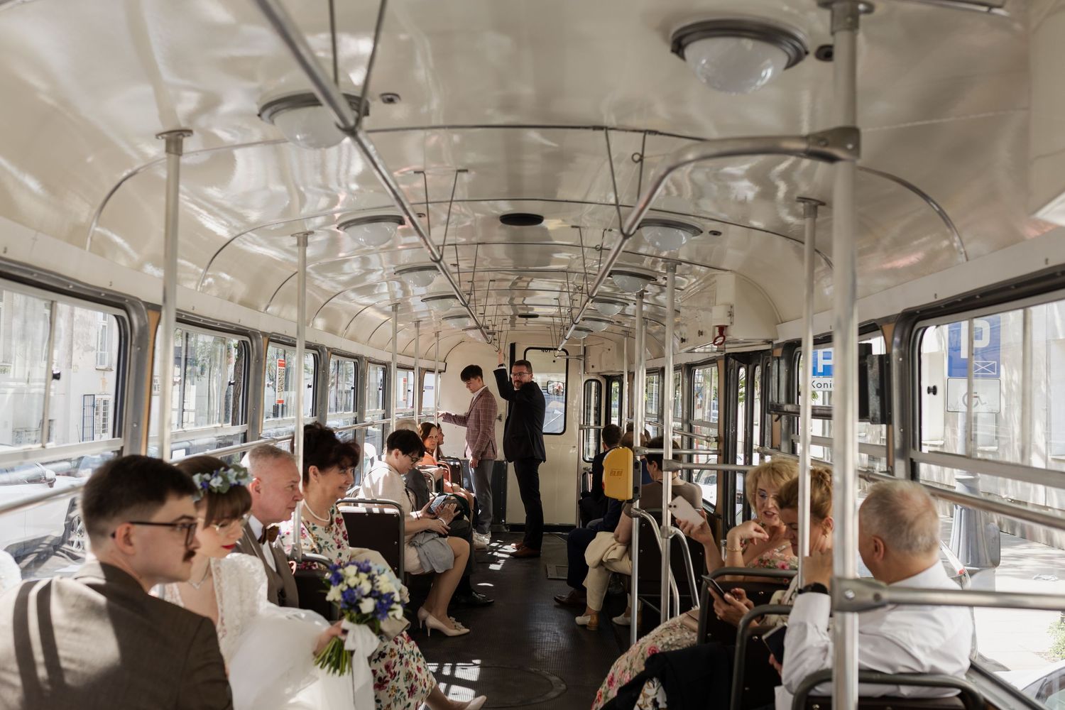 Interior view of a vintage tram car with passengers seated along both sides and standing during a daytime commute.