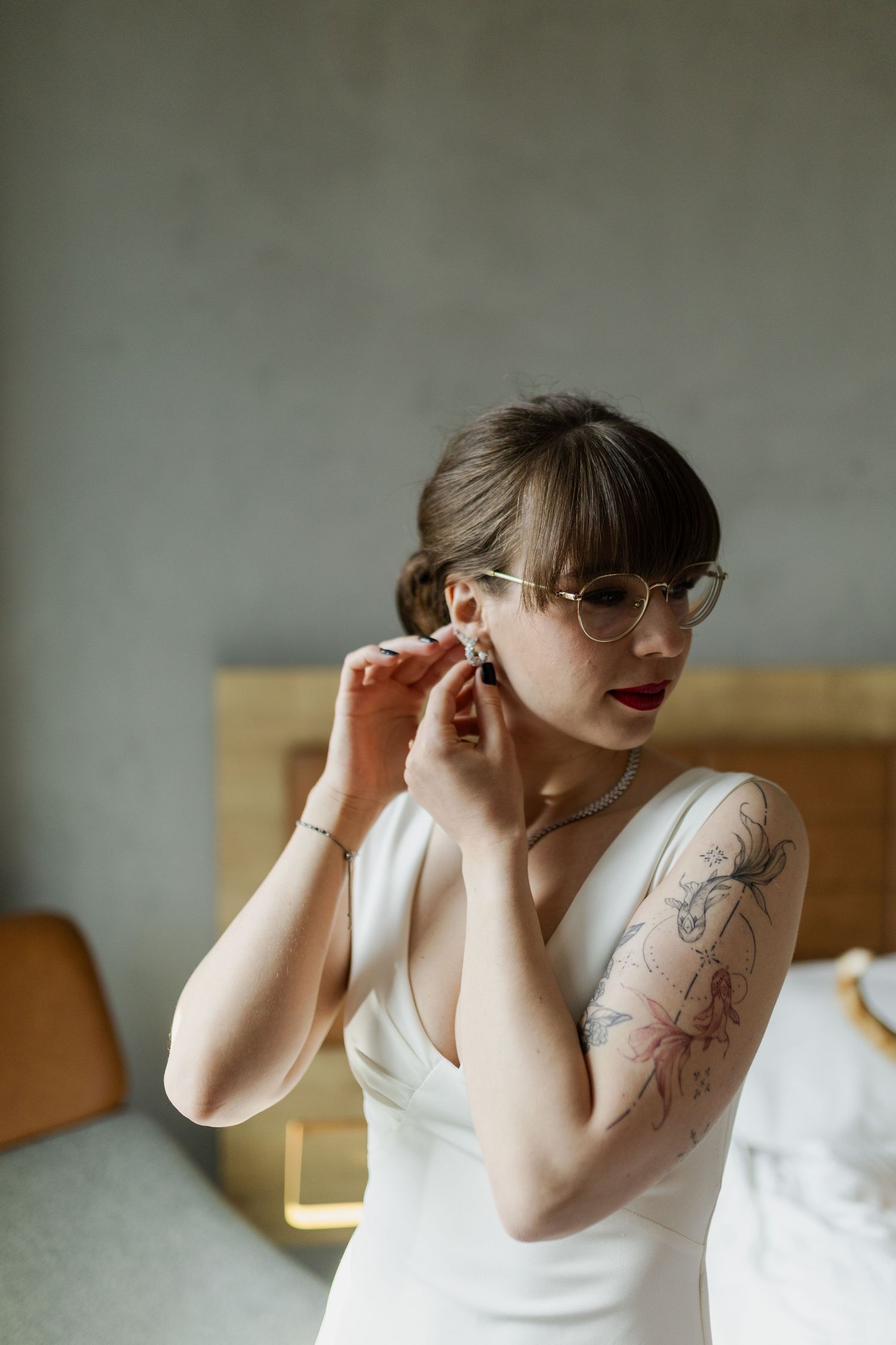 Someone wearing a white dress and glasses adjusts an earring, showing floral tattoo art on their arm.